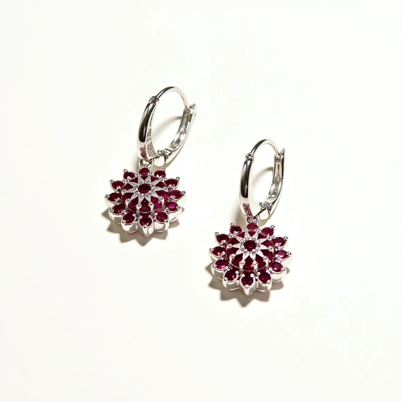 These floral earrings feature a captivating design with a cluster of vibrant red gemstones, likely rubies, arranged in a circular flower pattern. The stones appear to be round cut, securely set in prong settings that accentuate their brilliance. The earrings are crafted from a shiny silver-toned metal, possibly sterling silver or white gold, offering a polished and elegant look. The earrings are attached with a hinged hoop clasp, ensuring secure and comfortable wear, while the floral motif adds a touch of elegance and charm to the overall design.