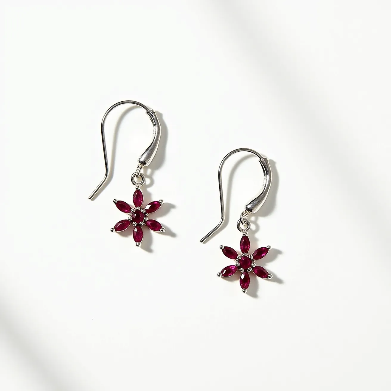 These floral earrings feature a delicate design with petals made from marquise-cut, deep red gemstones likely representing rubies or a similar stone. These gemstones are arranged in a symmetrical, flower-like pattern radiating from a central point. The stones are prong-set, which allows the vivid color of the gemstones to be prominently displayed. Crafted from a lustrous silver-toned metal, the earrings exude elegance and sophistication. The hook-style clasps provide a secure yet comfortable fit, seamlessly complementing the overall design while allowing the floral motif to dangle freely and catch light beautifully.