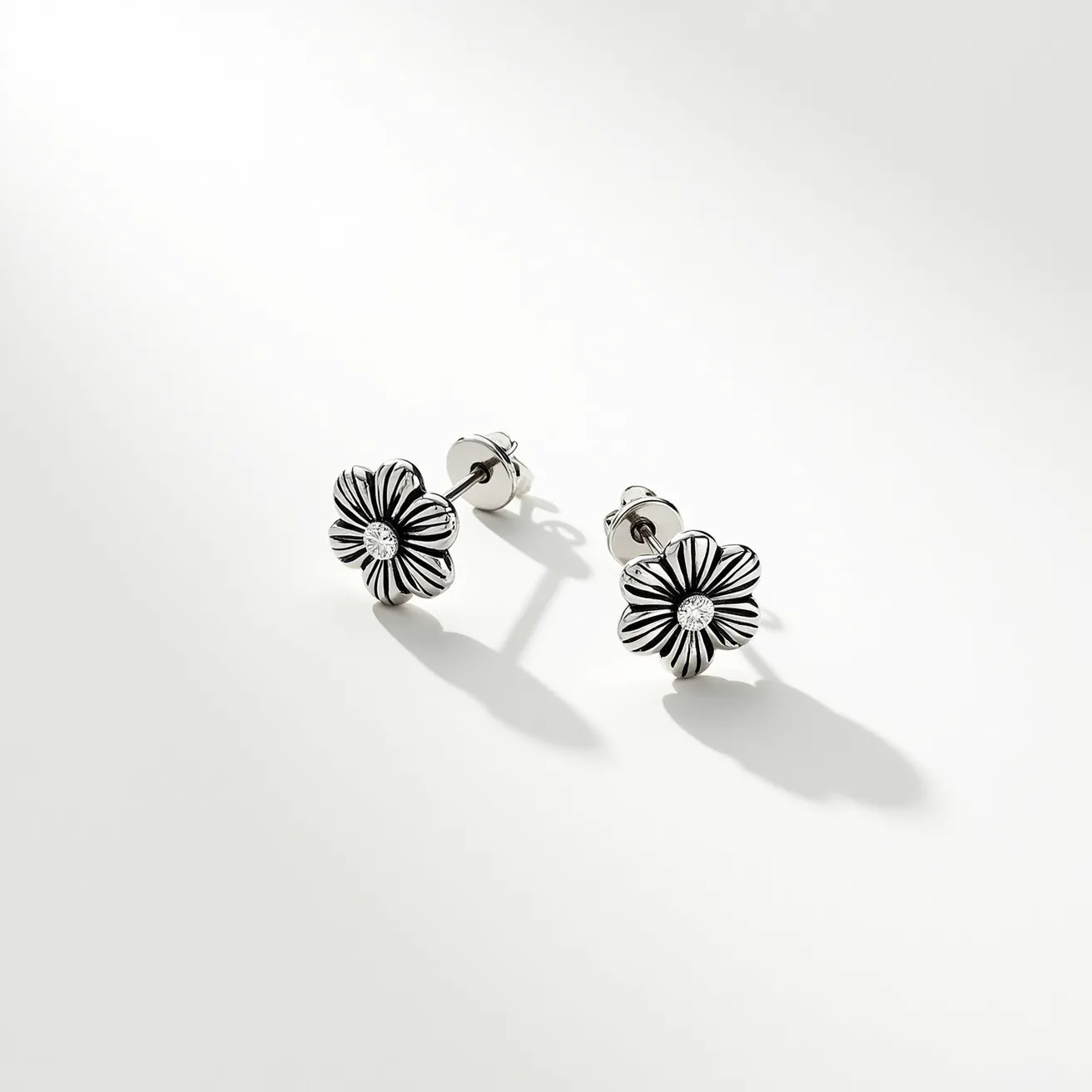 These floral earrings feature a charming design with intricately crafted petals made from what appears to be a polished metallic material, possibly silver, giving them a sleek and shiny appearance. At the center of each flower, there is a round, brilliant-cut gem that resembles a diamond, elegantly set to catch the light and add sparkling detail. The edges of the petals are defined with a darkened inlay, which adds depth and contrast to the floral motif. These earrings have a classic post-and-butterfly clasp, ensuring they remain secure when worn. The overall design showcases a perfect blend of fine craftsmanship and elegant simplicity.