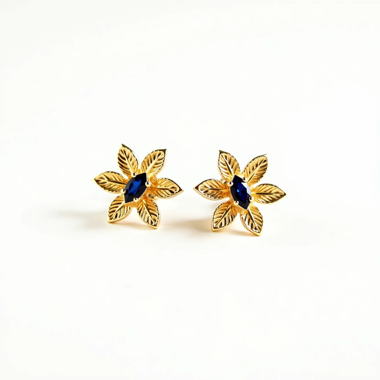 These floral earrings have a captivating design featuring a delicate arrangement of gold-toned petals, each with intricate detailing that resembles natural leaves. At the center of the flowers, a blue, marquise-cut gemstone is prong-set, adding a vibrant pop of color and elegance to the overall design. The earrings appear to utilize a stud attachment that secures them comfortably on the ears. The harmonious blend of warm gold and cool blue creates a striking contrast, making these earrings both versatile and eye-catching.