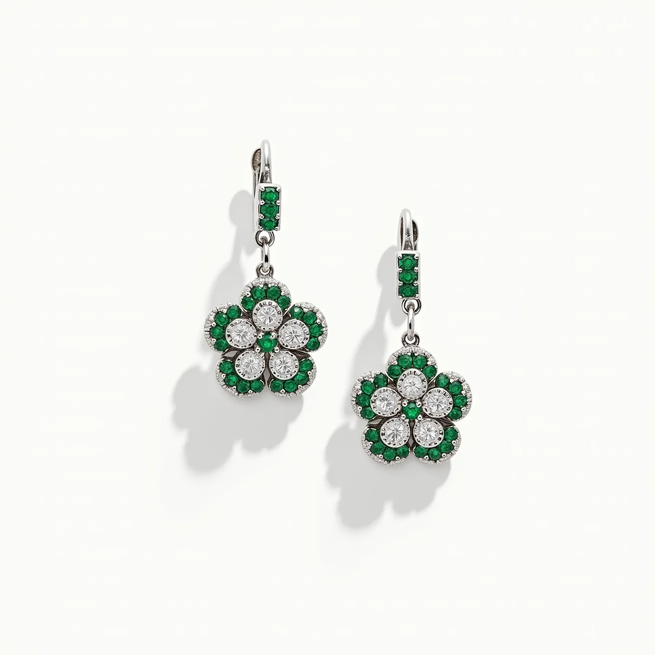 These flower earrings feature a delicate floral design with a combination of sparkling gems. Each earring displays a flower with petals formed by clustered circular-cut white stones, possibly diamonds, arranged around a central green stone, likely an emerald, which adds a vibrant contrast. The perimeter of each petal is lined with smaller green stones, enhancing the floral motif. The gems are securely set in what appears to be a white metal, possibly white gold or platinum, that provides a sleek, modern finish. The earrings are affixed with lever-back clasps, ensuring a secure and elegant attachment.