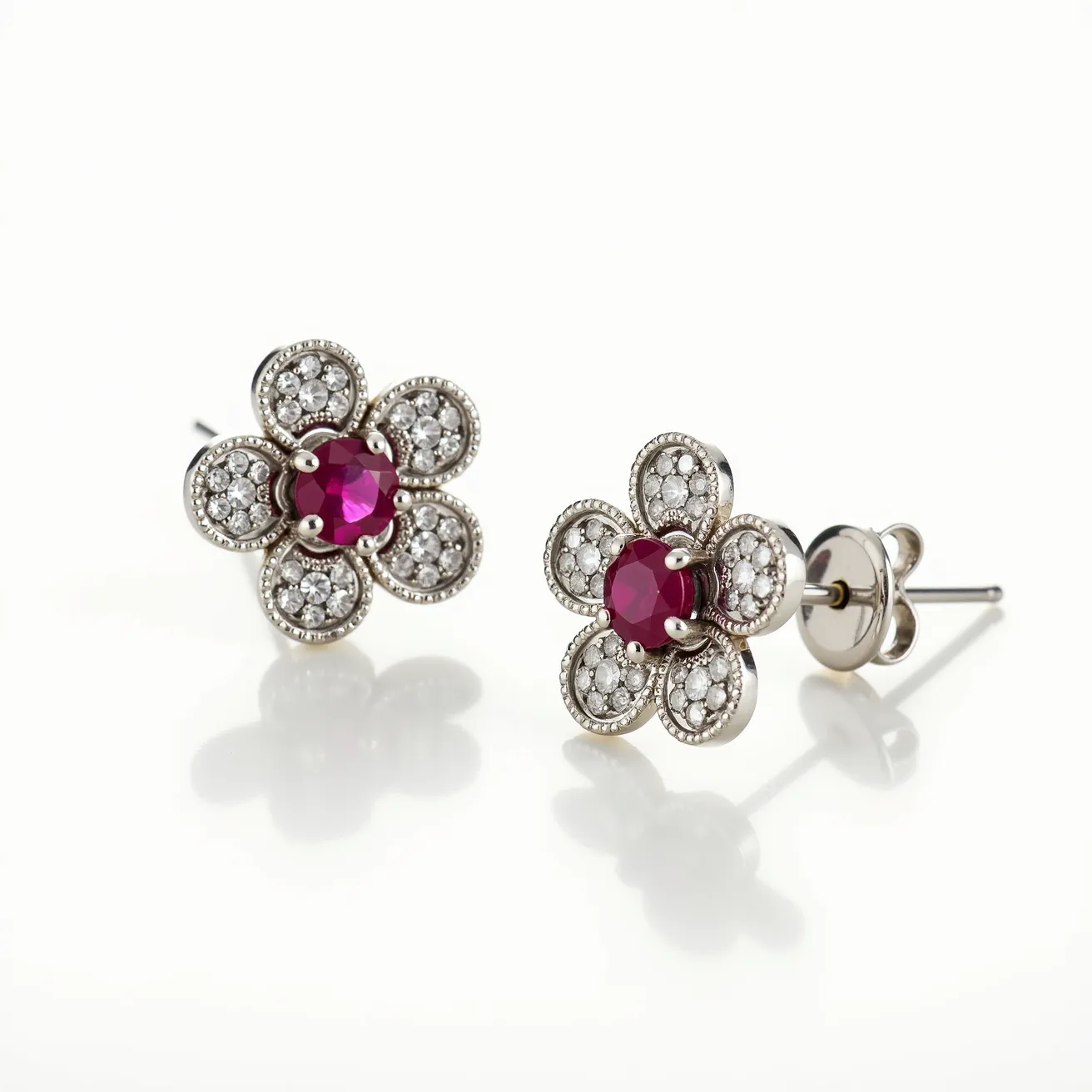 These flower earrings feature a charming design with each earring shaped like a delicate flower. The petals are adorned with clusters of small, round-cut diamonds set in pavé style, adding a sparkling allure. At the center of each flower is a striking ruby gemstone, cut in a brilliant round shape, secured by four prongs. The metal of the earrings appears to be polished white gold, enhancing the brilliance of the diamonds and the vivid color of the rubies. These earrings are designed with a post and friction back clasp mechanism, ensuring they stay securely in place while worn. Overall, the combination of rubies and diamonds in a floral motif creates a classic, elegant look.