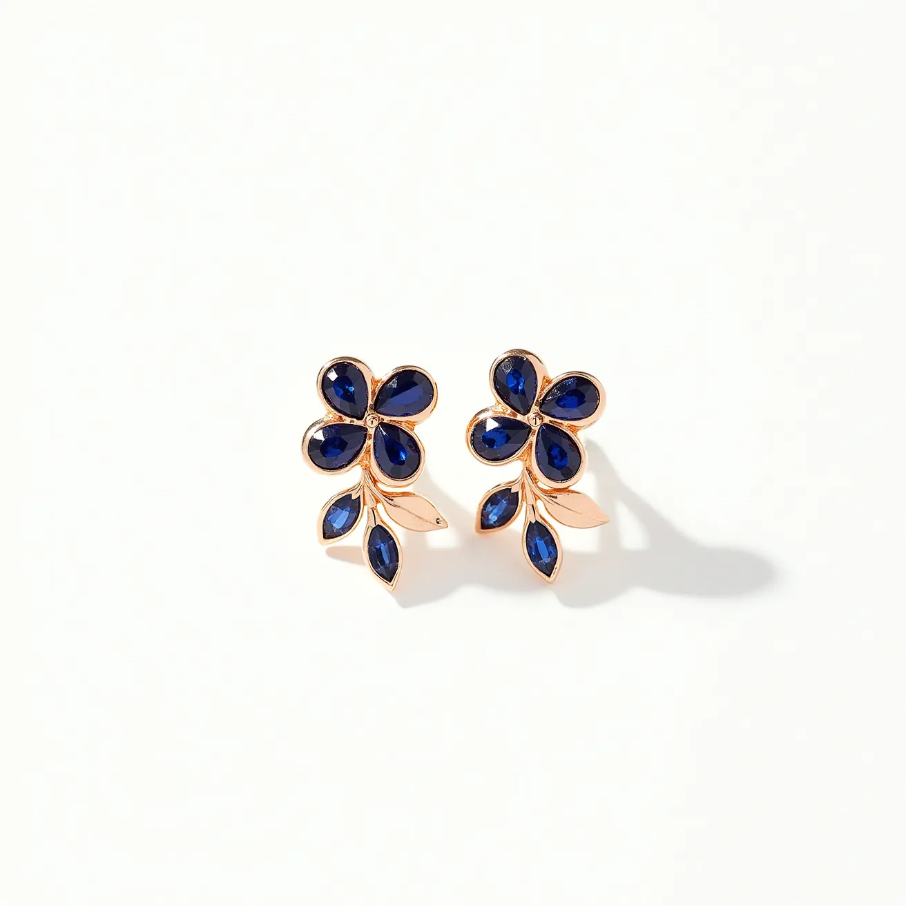 These flower earrings design features a delicate arrangement of petals and leaves, crafted from what appears to be a metal with a polished finish, possibly gold or a gold-toned material. The petals of the flowers and leaves are adorned with deep blue gemstones, likely sapphires or blue crystals, which are teardrop-shaped and set into the metal. These stones are faceted to enhance their sparkle and are arranged in a manner that mimics the structure of a flower with leaves beneath. The gems appear to be securely held in place with a bezel or similar setting, framed by the metal. A post and butterfly clasp attachment is visible, providing a secure fitting when worn.