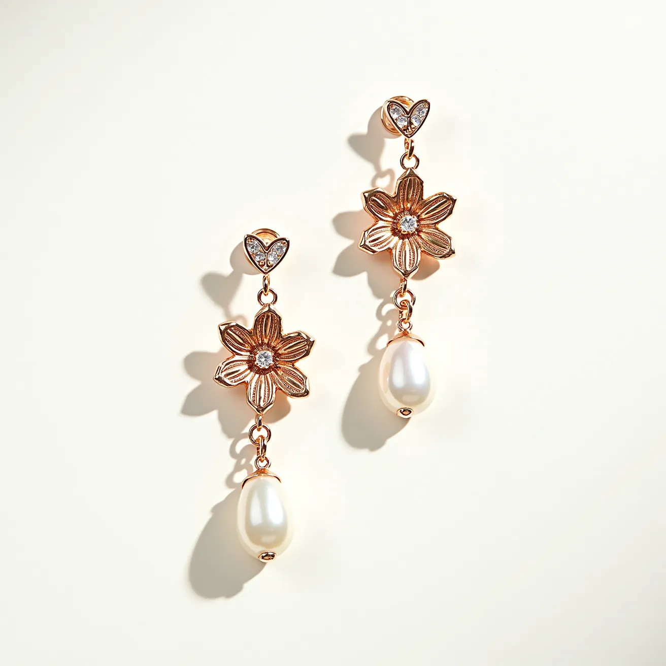 These flower earrings feature a delicate floral design crafted from a warm-toned metal, likely gold or a gold alloy. Each earring is adorned with a central gemstone set within the flower, appearing to be a round-cut diamond or crystal in a prong setting, enhancing the sparkling effect. Above the flower, a heart-shaped setting holds additional small round gems, adding a touch of elegance and intricacy. Dangling from each floral centerpiece is a lustrous teardrop-shaped pearl, smoothly polished to complement the overall design. The earrings are likely attached with a post-back or stud fastening, ensuring a secure hold while worn.