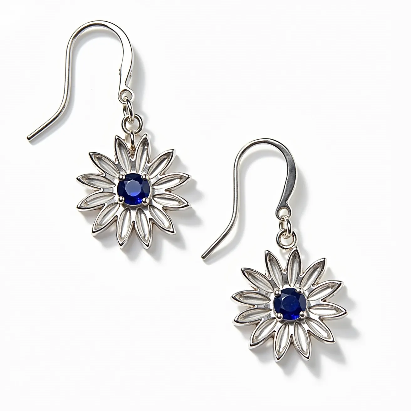 These flower earrings feature an intricate design crafted from a polished metal, likely silver, forming a floral motif with pointed petal details. At the center of each earring is a round, faceted blue gemstone, set in place with a classic prong setting that securely encases the stone while allowing light to enhance its deep hue. The earrings utilize a simple and elegant fishhook clasp for easy wear, adding to the overall delicate aesthetic of the piece. The combination of the cool-toned metal and vibrant stone creates a striking yet sophisticated accessory.