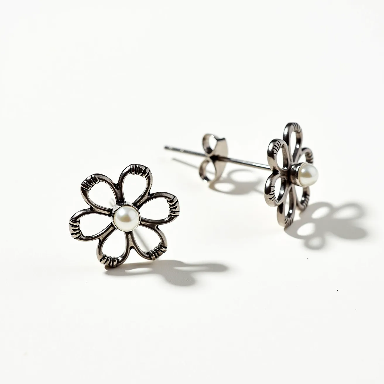 These flower earrings are crafted with a metallic material shaped into a delicate floral design. Each earring features a single, centrally placed round pearl, lending a touch of elegance and sophistication. The pearls appear to be set securely in a bezel setting, which enhances their prominence in the design. The earrings use a post and butterfly clutch attachment, a common and secure method for stud earrings, allowing for easy and comfortable wear. The intricate detail in the metalwork adds texture and depth, complementing the smooth surface of the pearls.