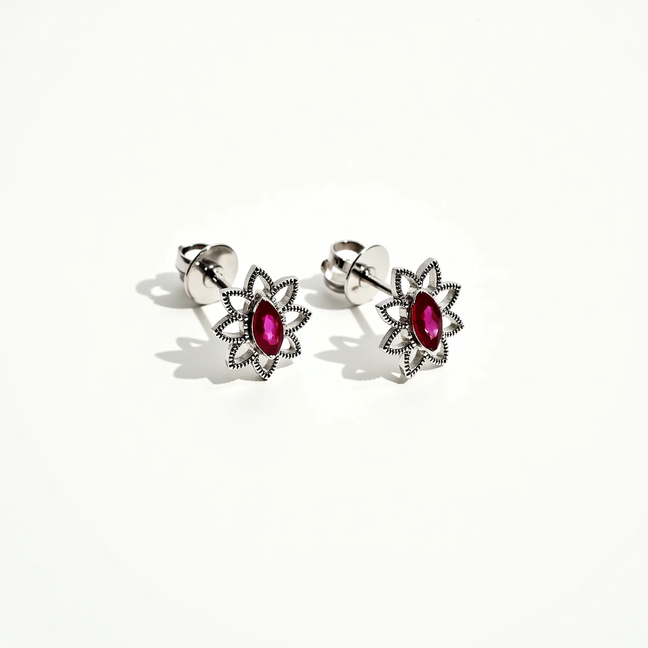 These flower earrings feature a delicate floral design crafted from a silver-toned metal, forming a dainty petal shape. At the center of each earring is a striking marquise-cut red gem, securely set in a prong setting that enhances its vibrancy and elegance. The earrings are equipped with a butterfly clasp attachment, providing a secure and comfortable fit. The intricate detailing around the petals adds a textured look, complementing the overall aesthetic of the piece.