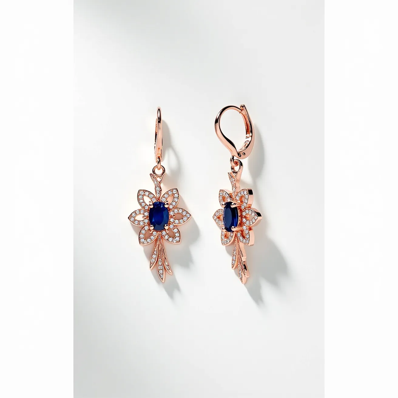 These flower earrings are crafted with a rose gold-toned metal, forming an intricate floral shape. At the center of each flower is a deep blue oval gem, likely a sapphire, securely set within a bezel or prong setting. Surrounding the central stone are numerous small, clear stones, possibly diamonds or cubic zirconia, highlighting the petals with a pave setting. The earrings are designed with a leverback clasp, ensuring a secure and elegant attachment to the ear. The rose gold color complements the blue stones, creating a striking and harmonious look.