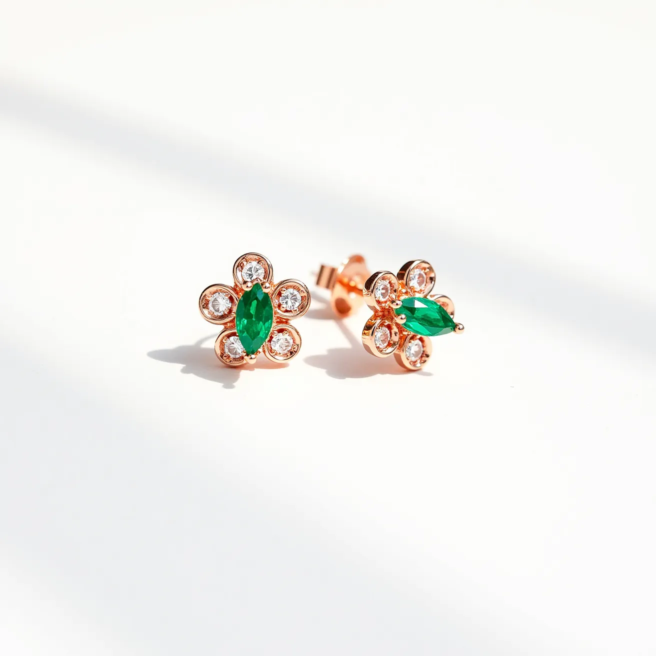 These floral stud earrings feature a delicate design crafted with a rose gold metal setting. Each earring has a central, vividly green marquise-cut gemstone, possibly an emerald, which serves as the focal point. Surrounding the center stone are small round brilliant-cut clear gemstones, likely diamonds or cubic zirconia, mimicking the petals of a flower. The gems are securely set in prong settings to ensure durability. The earrings are attached with a post and butterfly clasp, offering secure and comfortable wear.