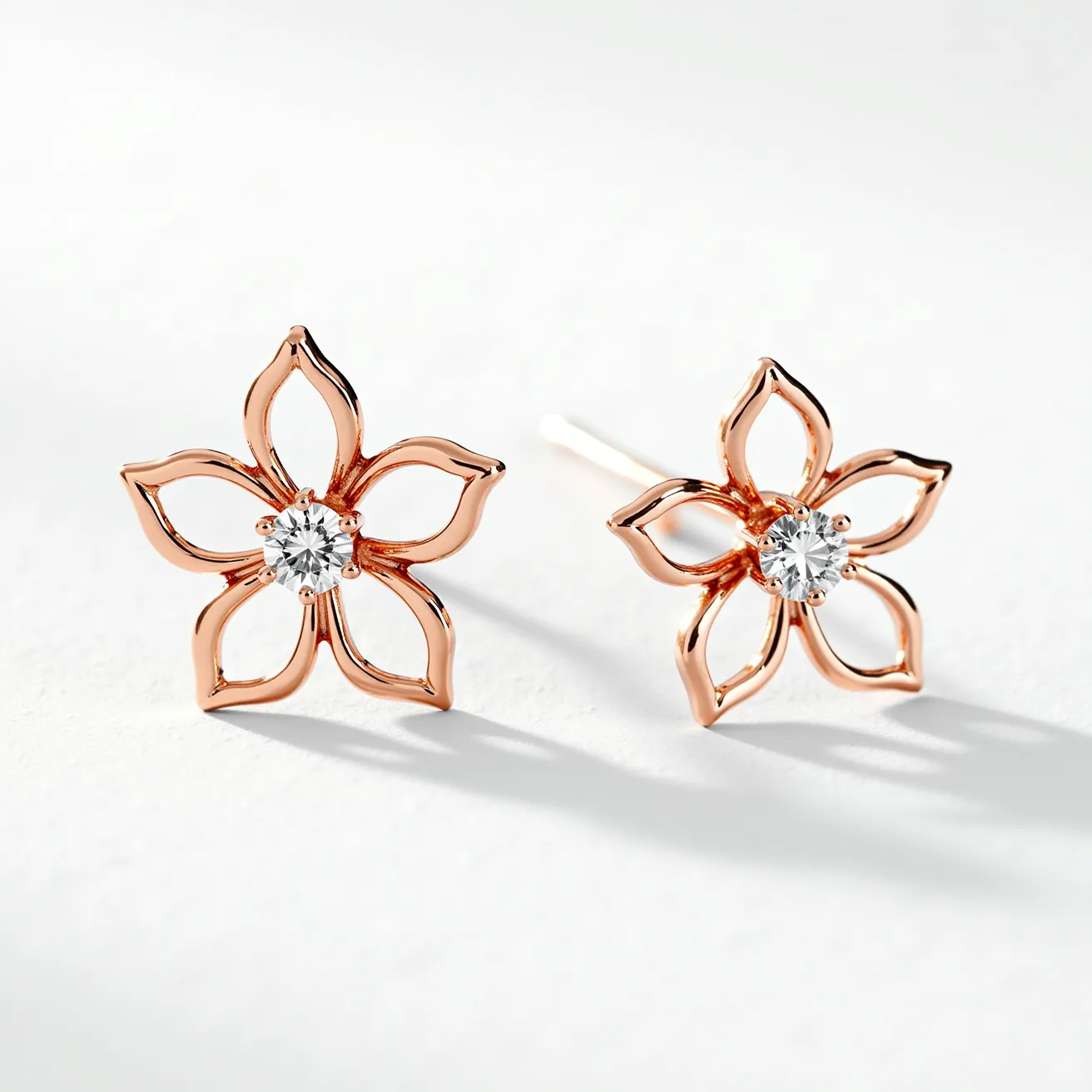 These floral stud earrings feature an elegant design crafted from rose gold, shaped into delicate, open-petaled flowers. At the center of each flower is a round-cut white gemstone, likely a diamond, securely held in place by a classic prong setting. The stud earrings have a sleek, straight post which suggests a secure push-back clasp is used for attachment, ensuring both style and functionality. The combination of shimmering gemstones and the warm hue of rose gold gives these earrings a sophisticated and timeless appeal.