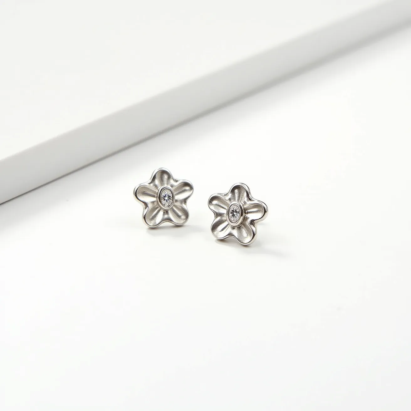 These floral stud earrings feature a delicate flower design crafted from a shiny metallic material that resembles polished silver or white gold. At the center of each earring is a small, round-cut clear gem, possibly a diamond or cubic zirconia, held securely in place with a bezel setting. The earrings are designed with a simple post attachment, making them easy to wear and secure with a standard push-back or butterfly clasp, which is ideal for stud earrings. The overall aesthetic is elegant and minimalist, suitable for both everyday wear and special occasions.