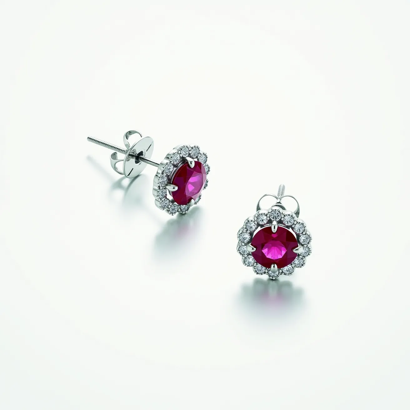 These floral stud earrings feature a vibrant, round-cut red gemstone at the center, likely a ruby, showcasing a deep, captivating hue. The central stone is encircled by a halo of sparkling, round-cut white diamonds, each meticulously set in a prong setting to form a floral pattern that enhances the overall brilliance and elegance of the design. The earrings are crafted from a lustrous silver or white gold metal, which adds a cool, modern contrast to the vivid red center and the shimmering diamonds. The attachment is a secure screw-back post, ensuring a comfortable and stable fit for all-day wear.