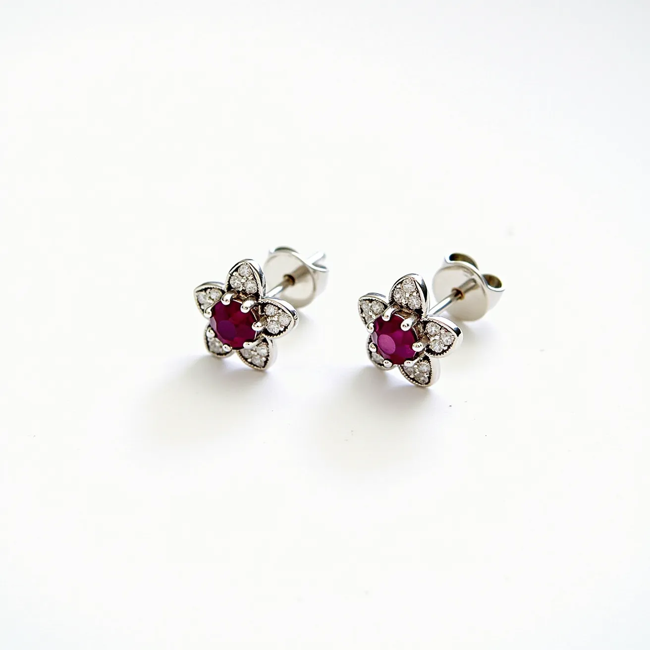 These floral stud earrings feature a striking design centered around a vibrant red gemstone, likely a ruby, set in a secure prong setting. Surrounding the central stone are petal-shaped arrangements of small, sparkling clear gemstones, possibly diamonds or cubic zirconia, creating a floral motif. The earrings are crafted from a shiny metallic material, likely silver or white gold. They utilize a post and butterfly clutch fastening, ensuring they stay securely in place when worn. The combination of the rich red center stone and the shimmering clear stones gives these earrings an elegant and timeless appeal.