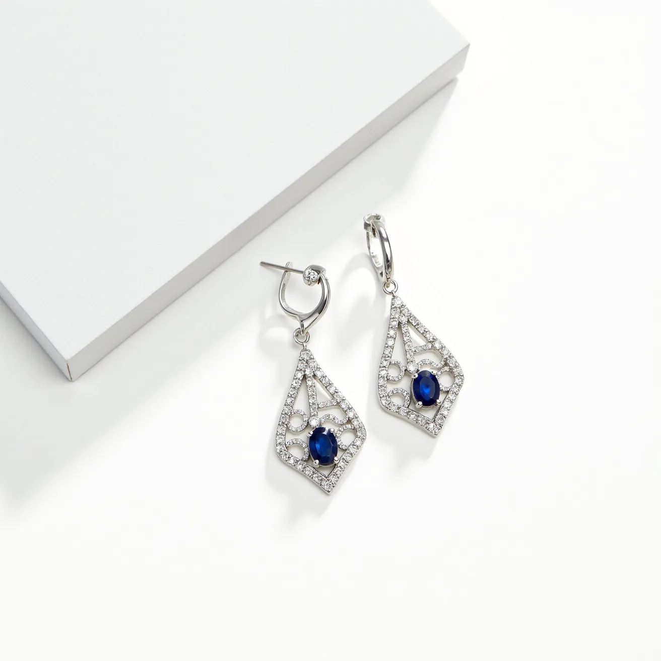 These gemstone earrings feature an intricate design with a prominent oval blue sapphire at the center, elegantly surrounded by a series of small, round cut clear gemstones, likely diamonds. The stones are set within a delicate latticework of silver or white gold, forming an ornate and symmetrical geometric shape. The earrings are attached to the ear with a secure hoop clasp, through which the main pendant dangles, allowing the design to sway gently with movement. The overall look is one of sophistication and timeless elegance, showcasing the craftsmanship and the harmonious blend of materials used.