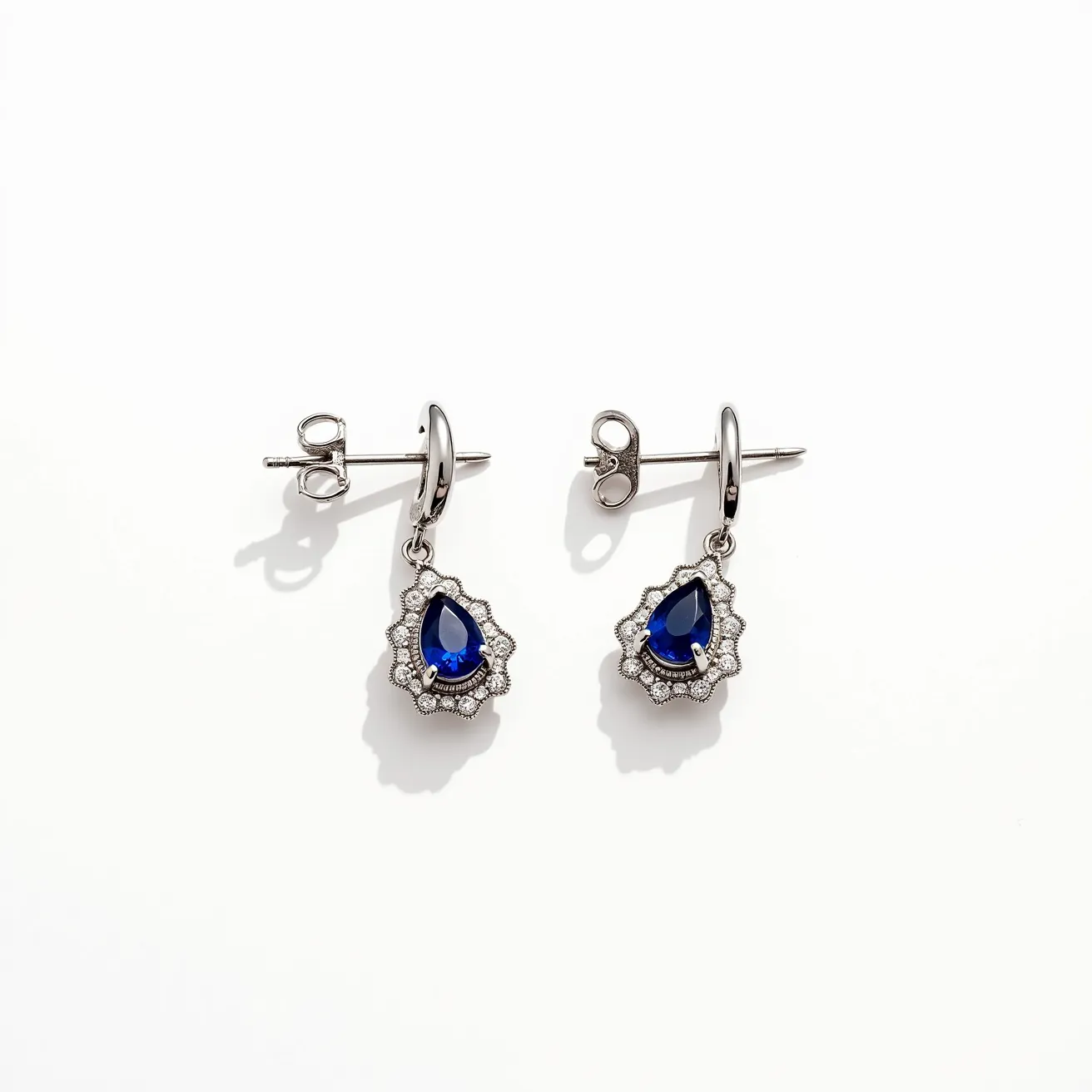 These gemstone earrings feature elegant teardrop-shaped blue gemstones, likely sapphires, set in a polished metal that appears to be silver or white gold. Surrounding the central stones, a halo of small clear stones, potentially diamonds or cubic zirconia, enhances their brilliance and adds a classic touch. The earrings are designed with a small hoop that attaches to a post and a butterfly clasp, ensuring secure wear. The meticulous arrangement and quality of the gems together create a timeless and sophisticated appearance, showcasing a blend of vibrant color and refined sparkle.