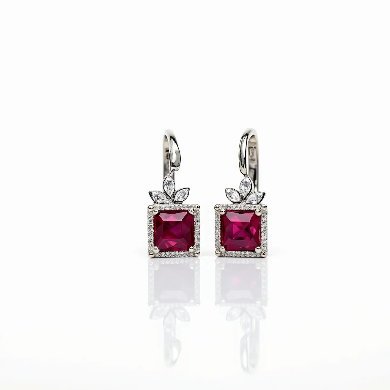 These gemstone earrings feature a striking design centered around vivid, square-cut red gemstones, elegantly set in a prong setting. The gemstones are framed by a series of small, brilliant-cut clear stones creating a halo effect. Above each red stone, there are additional leaf-shaped clear stones, possibly diamonds, arranged in a floral motif. The earrings are crafted from a shiny metal, likely white gold or platinum, which enhances the vibrant color of the gemstones. They are fitted with a leverback clasp that ensures secure and comfortable fastening.