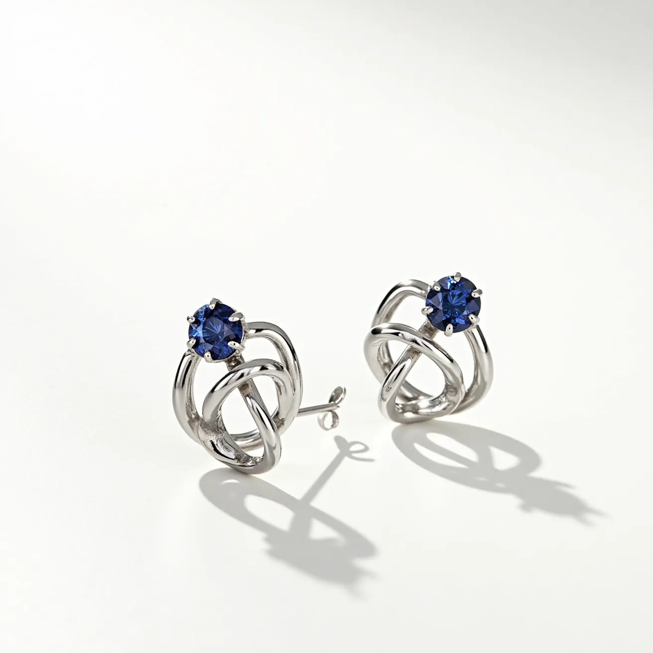 These gemstone earrings feature a pair of deep blue sapphires, each set in a prong setting that securely holds the stones in place. The sapphires are cut in a round shape, which enhances their brilliance and depth of color. The design of the earrings includes an intricate metalwork in a swirling pattern, possibly made of white gold or platinum, which adds elegance and sophistication to the overall look. The earrings are equipped with a post and butterfly clasp, ensuring a secure and comfortable fit for the wearer.