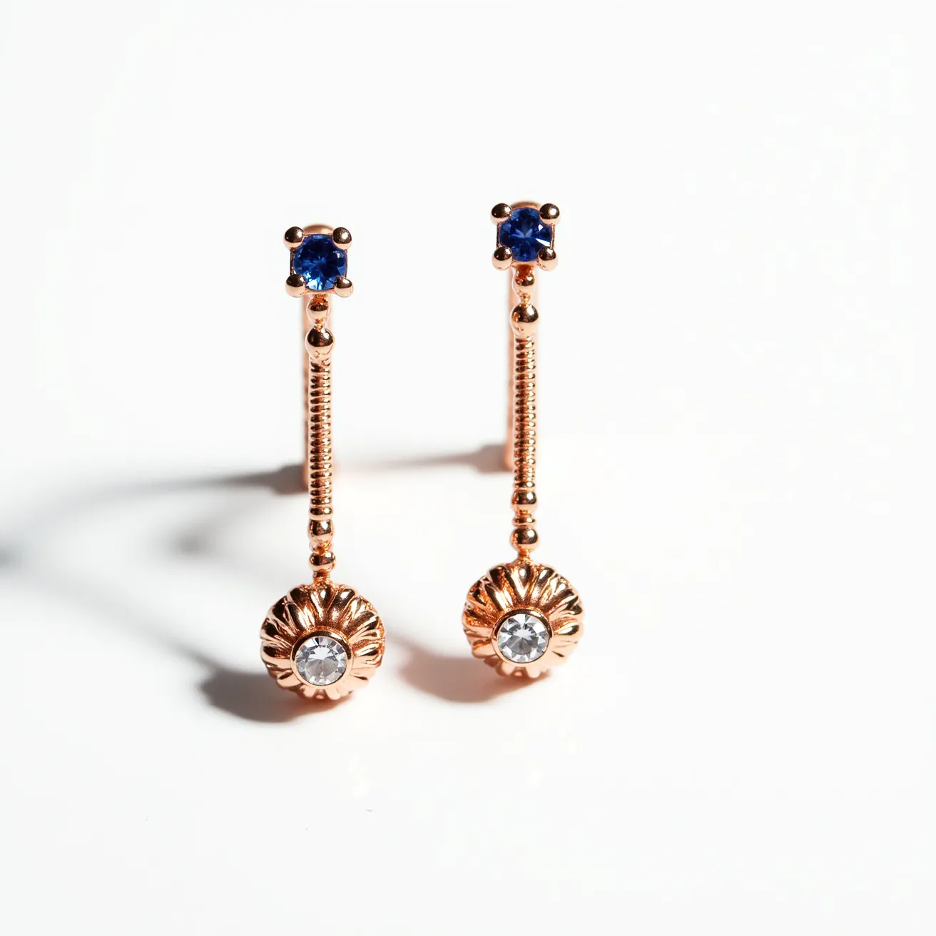 These gold cartilage earrings feature an elegant design with a vertical bar encrusted with a small blue gemstone at the top, set in a classic prong setting. The blue gemstones are round in shape, adding a subtle hint of color to the piece. At the end of the bar hangs a floral-like pendant with a central clear gemstone, also round, and secured within a bezel-like setting, offering a distinctive touch. The use of gold complements the stones beautifully, enhancing the luxurious feel of the earrings. The clasp design is not visible, but the earrings are crafted to sit comfortably on the ear cartilage.