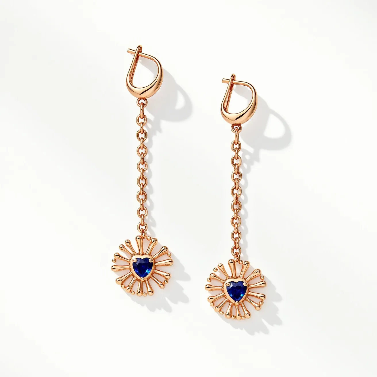 These gold dangle earrings feature a delicate chain design that suspends a radiant sunburst motif, each centered with a deep blue heart-shaped gemstone. The stones are set securely in a bezel setting, enhancing their vibrant color and adding a touch of elegance to the overall design. The earrings are crafted from gold, which complements the rich hue of the gemstones beautifully. They include a hoop clasp attachment, ensuring a secure and comfortable fit when worn. The intricate craftsmanship and striking combination of gold with the blue stones make these earrings a standout accessory for various occasions.