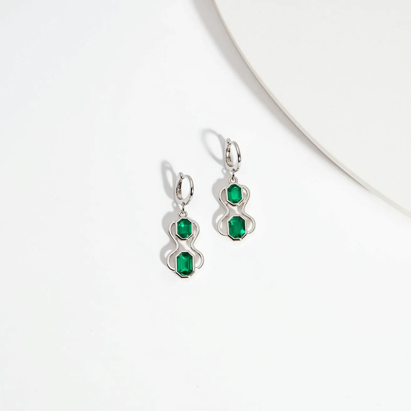 These gold dangle earrings feature a striking design with a sleek, elegant form. Each earring showcases two vibrant, rectangular-cut green gemstones, likely emeralds, set within a fluid, modern gold frame. The stones are secured in a prong setting, enhancing their brilliance and allowing light to pass through. The earrings attach with hoop clasps, ensuring a secure and comfortable fit. The combination of the luminous gold and the vivid green gemstones creates a luxurious and sophisticated look.