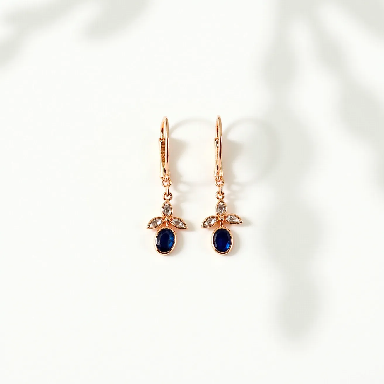 These gold dangle earrings feature an elegant design with a warm gold metal forming the primary structure. They are adorned with deep blue oval-cut gems, possibly sapphires, each set in a bezel that securely holds the stones in place. Accompanying these central gems are smaller, clear stones, potentially diamonds, expertly arranged in a leaf-like formation above the blue gems. The setting of these smaller stones appears to be prong-set, allowing for a classic, secure display. The earrings attach via a lever-back clasp, ensuring ease of wear while maintaining a refined aesthetic.