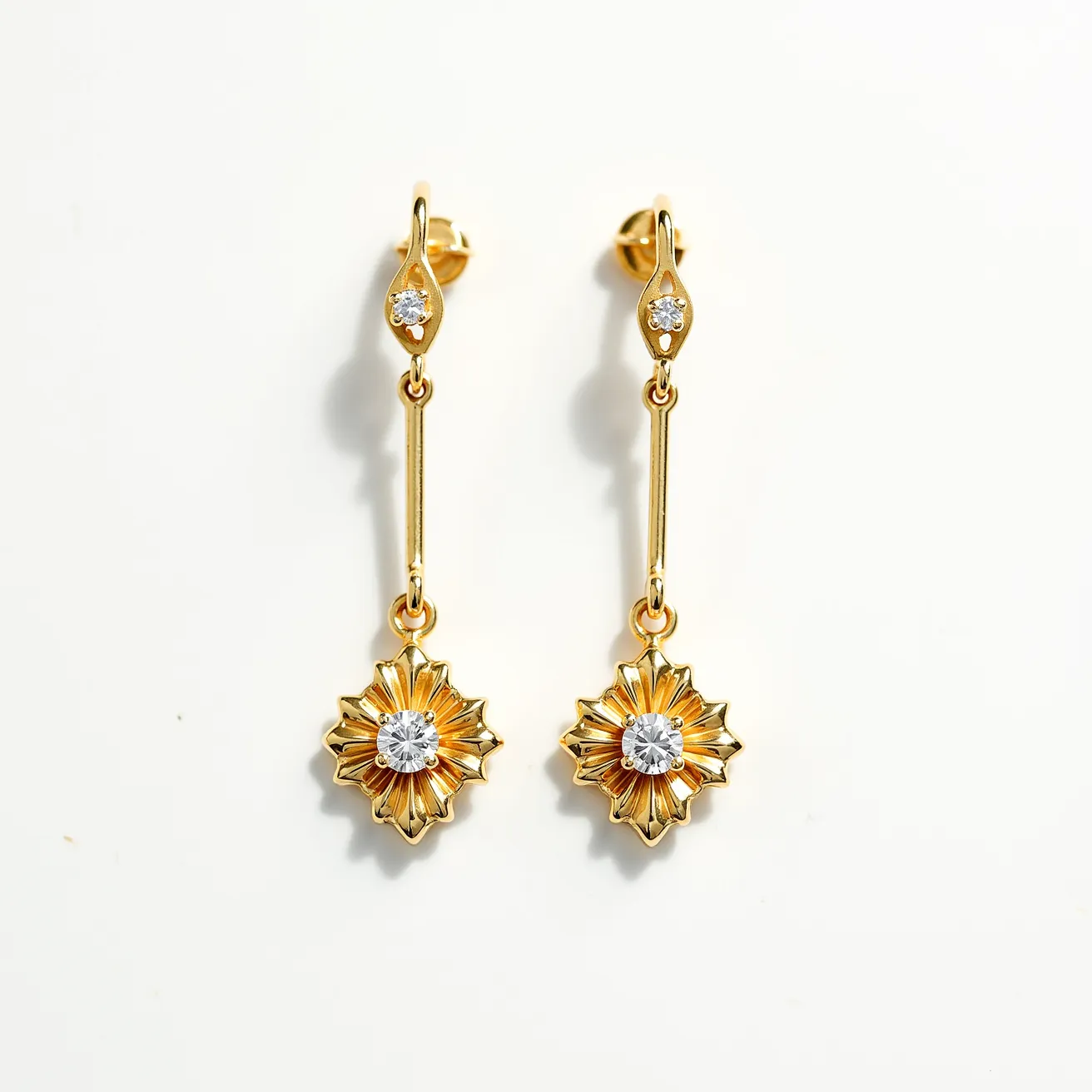 These gold drop earrings feature an elegant design with a prominent use of gold, creating a luxurious appearance. The earrings are adorned with sparkling round-cut gems at both the top and in the center of the starburst pendant, likely set in a secure bezel or prong setting, enhancing their brilliance. The drop design is elongated by a slender gold bar, which connects the top gem setting to the starburst motif. The earrings are equipped with a post-back clasp, ensuring they are securely fastened when worn. The combination of the gold with the clear sparkling gems gives these earrings a classic yet striking appeal.