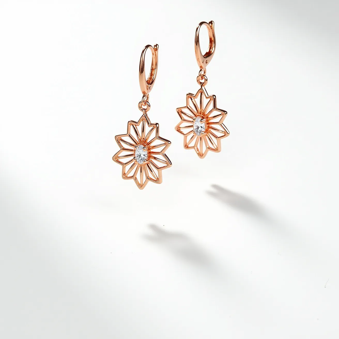 These gold drop earrings feature a radiant floral design, crafted from a rose gold metal. At the center of each flower is a small, oval-cut clear gemstone, securely held in place with a prong setting, adding a touch of sparkle to the overall aesthetic. The earrings are hinged with a hoop clasp, ensuring they hang elegantly and are securely fastened when worn.