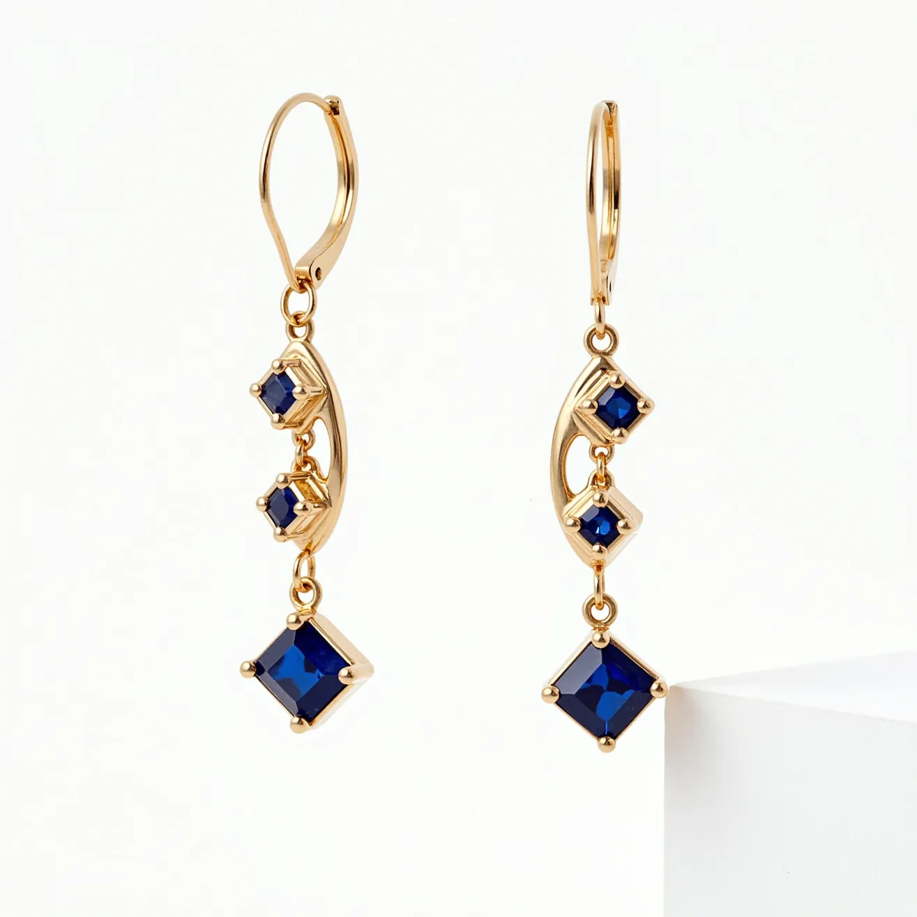 These gold drop earrings feature a delicate design crafted from polished gold, highlighted by elegant blue gemstones. The stones are square-cut and securely set in a prong setting, providing a striking contrast against the gold. The earrings are embellished with three gems each, positioned along a gracefully curved structure that enhances their sophisticated appeal. They are designed with a lever-back clasp, ensuring both security and comfort when worn.