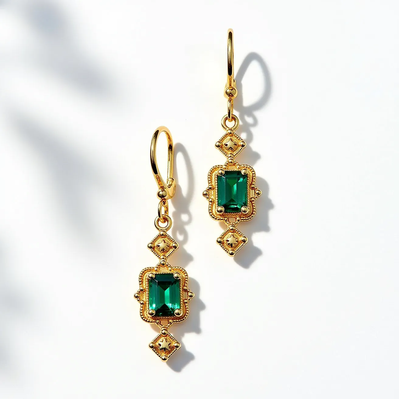 These gold drop earrings showcase a remarkable design featuring a polished gold base. The central element of each earring is a rectangular, emerald-colored gemstone set within an intricately detailed bezel setting, highlighting the vivid color of the stone. Above and below the main gemstone, there are small, diamond-shaped gold elements adorned with petite, clear stones, likely diamonds or cubic zirconia, adding a touch of sparkle and complexity to the design. The earrings have a secure hook clasp attachment, which complements the overall elegance and ensures comfortable wear.