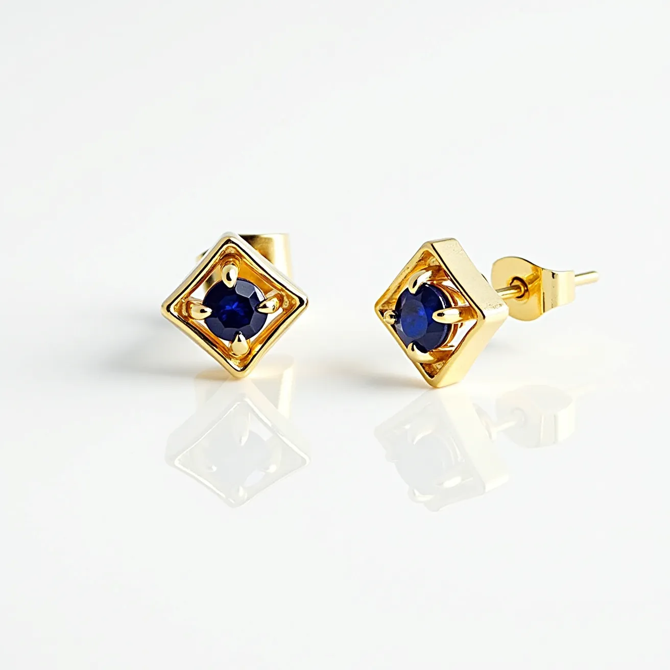 These gold earrings feature a geometric design with a central blue gemstone, likely a sapphire, set into a square gold frame. The stones are round-cut, enhancing their reflective qualities, and are securely held in place by four prongs. The earrings utilize a post and butterfly clasp for attachment, ensuring a secure fit when worn. The elegant combination of the rich blue stones and the warm gold setting creates a sophisticated and timeless look.