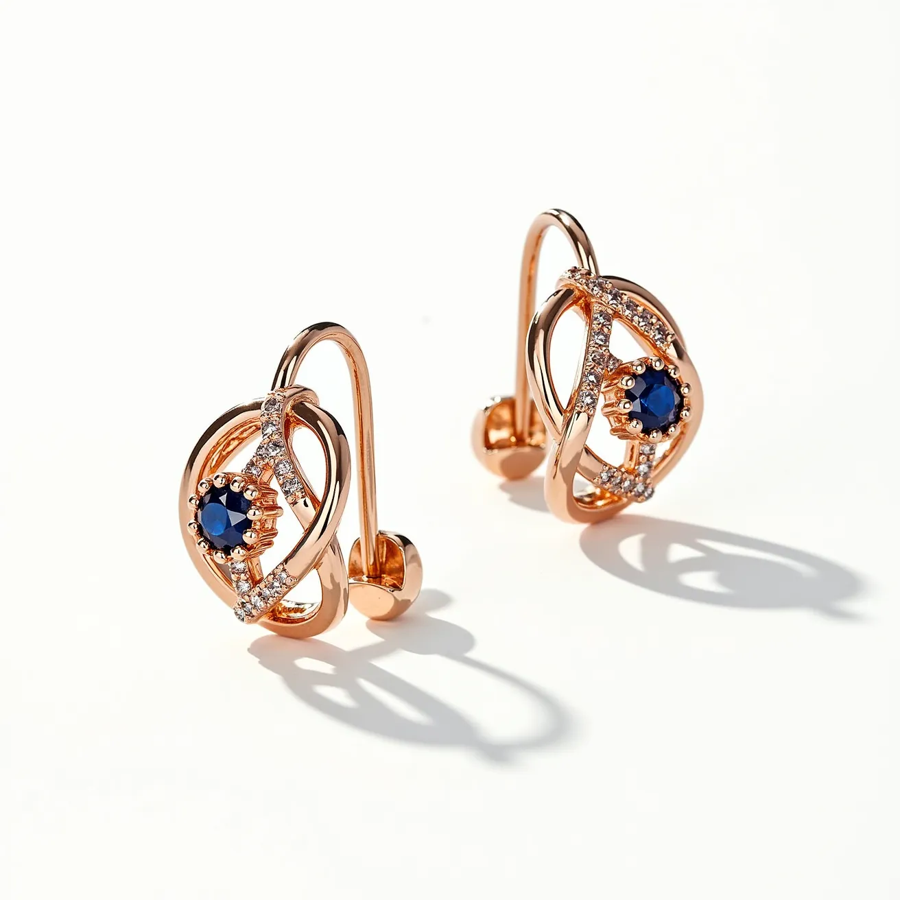 These gold earrings showcase an elegant design featuring an intertwined motif with a central deep blue gemstone, likely a sapphire, in a round cut. The setting is a prong type, which securely holds the gemstone in place while enhancing its visibility. Surrounding the central stone, small clear stones, presumably diamonds, are pave-set, adding a sparkling contrast to the blue gem. The gold used is rose gold, lending a warm and sophisticated tone to the overall appearance. The earrings are secured with a latch-back clasp, providing both comfort and security for the wearer.