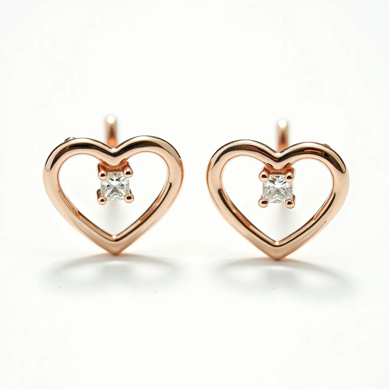 These gold heart earrings feature an elegant open-heart design crafted from what appears to be gold, possibly rose gold, giving them a warm and luxurious appearance. Each earring is adorned with a centrally set single clear gemstone, which is likely a diamond. The stone is cut into a square or princess shape and is securely held in place by a four-prong setting, adding a touch of sparkle and sophistication. The earrings are designed as studs, featuring a straight post for easy attachment to the ear, typically secured with a back clasp. The overall design is simple yet charming, making them suitable for various occasions.