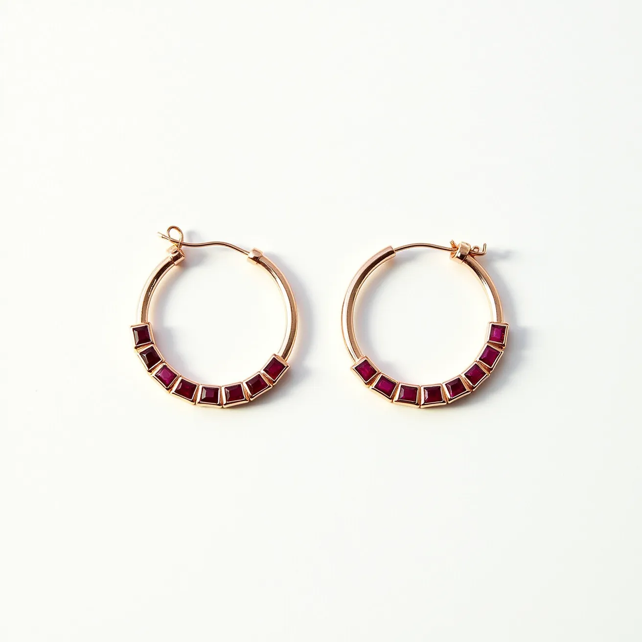 These gold hoop earrings feature an elegant circular design adorned with square-cut red stones, likely rubies, set in a bezel setting along the lower half of the hoop. The metal appears to be polished gold, providing a luxurious framework that complements the vibrant stones. They are equipped with a hinged wire clasp, ensuring a secure and stylish attachment to the ear. The craftsmanship highlights the contrast between the warm gold and the deep red stones, making these earrings a standout accessory.