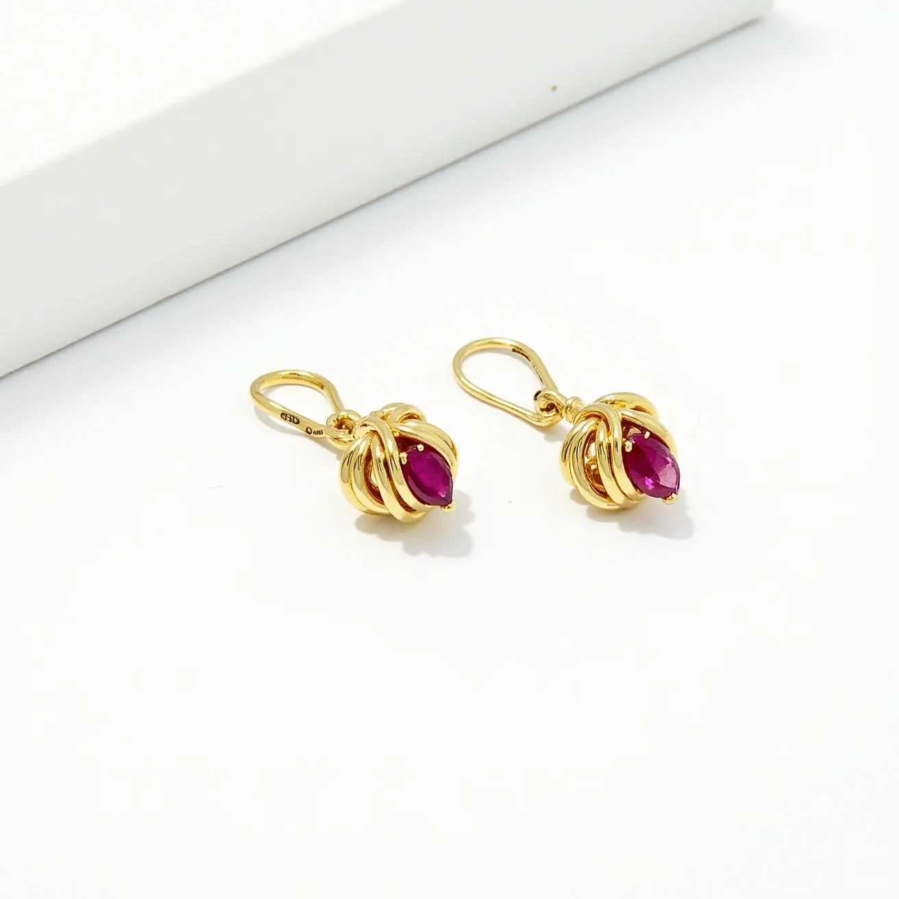 These gold knot earrings feature an elegant design crafted from polished yellow gold, showcasing intricate loops that form a classic knot shape. Centrally set within the knot is a vibrant oval-cut gemstone, likely a ruby, which is held securely in a prong setting. The rich red hue of the stone adds a striking contrast to the gold, enhancing its visual appeal. Each earring is attached with a leverback clasp, ensuring a secure and comfortable fit for the wearer.