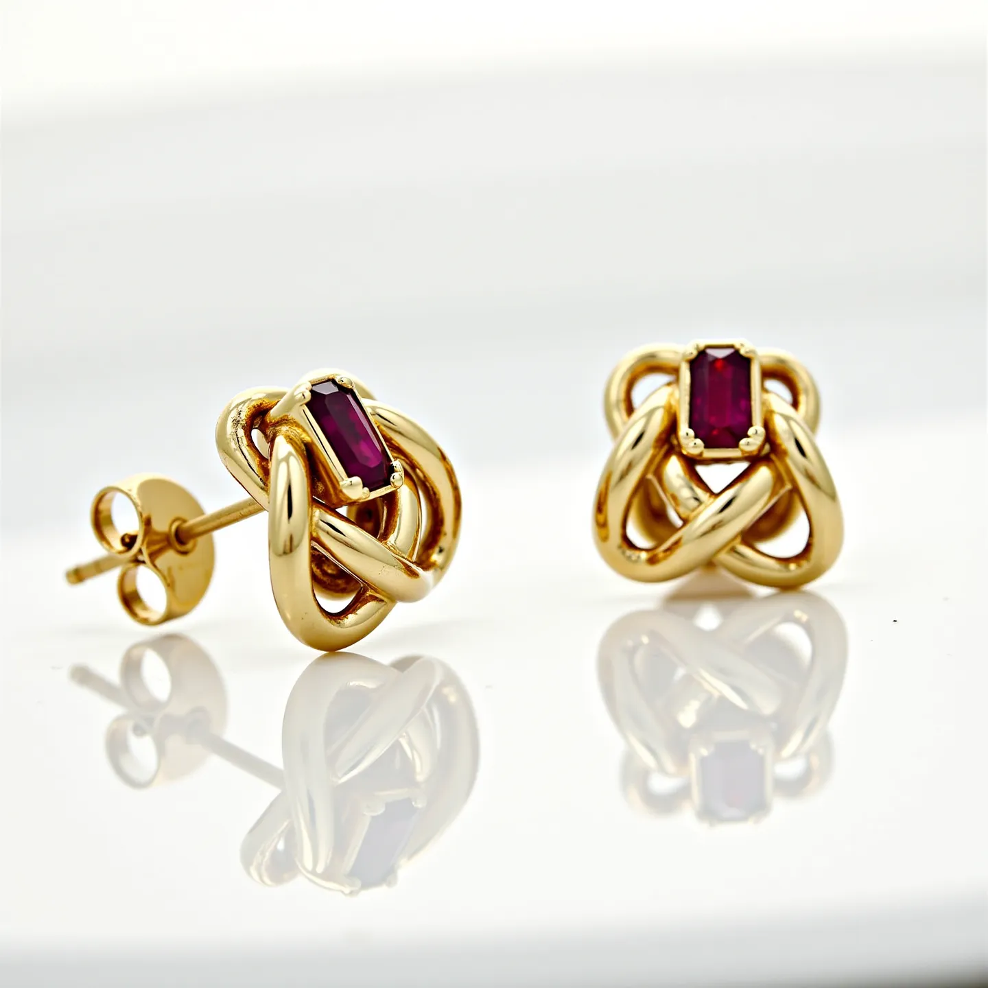 These gold knot earrings feature a beautifully intertwined design made of polished gold, giving them an elegant and sophisticated appearance. At the center of each earring is a striking, rectangular cut gemstone, possibly a ruby, held securely by a four-prong setting. The rich red hue of the gemstones adds a vibrant contrast to the warm gold tones of the knot design. These earrings use a post and butterfly back clasp, providing a secure fit when worn. The combination of the intricate knot motif and the vivid gemstones make these earrings a captivating accessory.