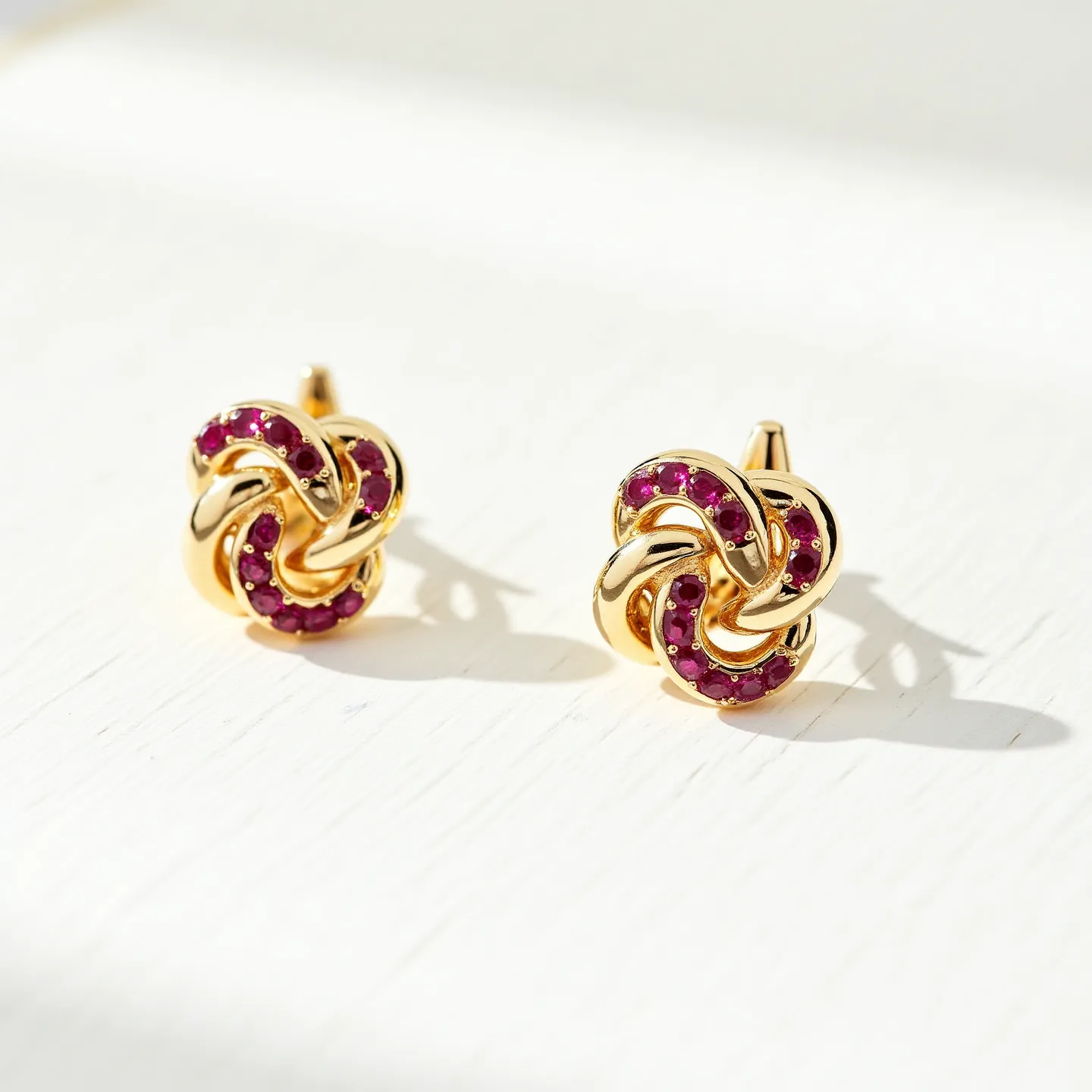 These gold knot earrings are crafted from polished gold, intricately designed into a knot shape that exudes elegance and sophistication. The earrings are adorned with small, round-cut stones that are likely rubies, set in a channel style along the curved strands of the knot, adding a vibrant touch of color. The back of each earring features a classic post, suggesting a secure push-back clasp mechanism to ensure ease of wear and stability. The harmonious combination of gold and gemstones makes these earrings a timeless and versatile accessory for various occasions.