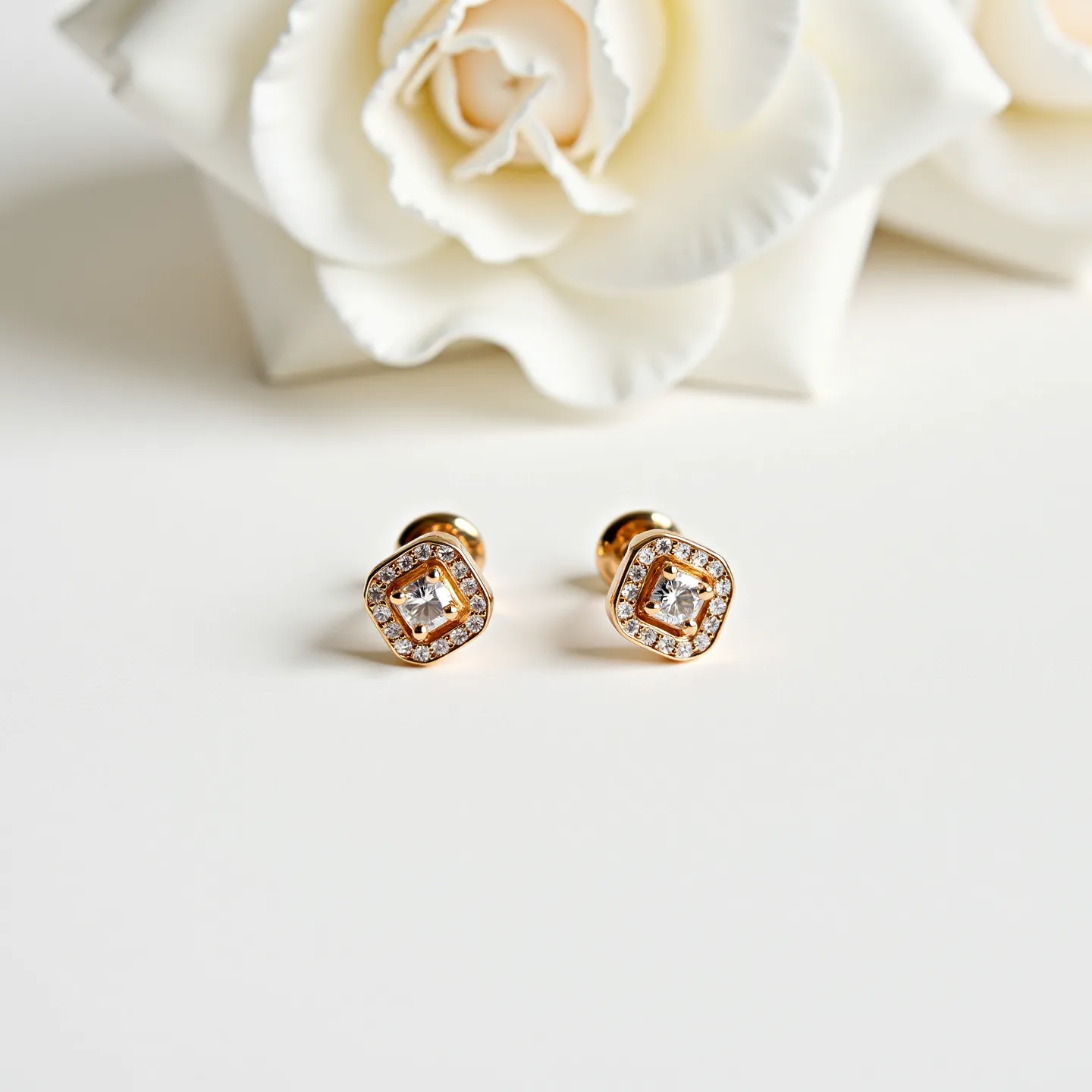 These gold stud earrings feature a central square-cut gemstone, likely a diamond given its reflective quality, encased in a rim of smaller, round-cut stones that enhance its brilliance. The main gems are meticulously set in a bezel setting that provides a secure fit while maintaining a sleek and modern aesthetic. The gold framework elegantly encircles the stones, contributing a warm and luxurious tone to the overall design. These earrings are attached with a push-back or butterfly clasp, a classic choice that ensures comfort and security for everyday wear. The combination of high-quality materials and thoughtful design details makes these earrings an exquisite addition to any jewelry collection.