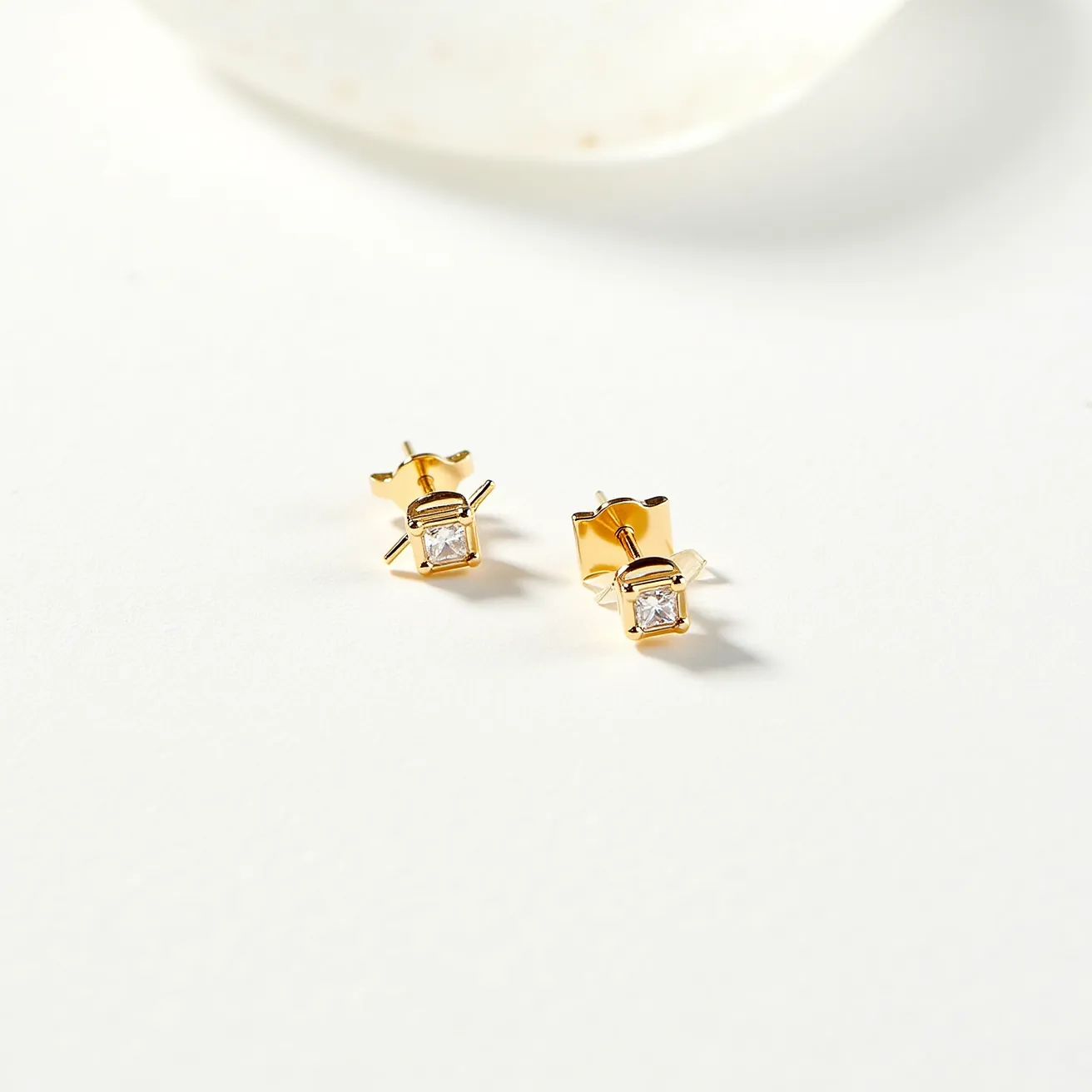 These gold stud earrings feature a simple yet elegant design crafted from gold metal. Each earring showcases a single, square-cut gemstone, likely a diamond or diamond simulant, securely set in a bezel setting that complements the gold’s warm luster. The earrings are fastened with a traditional post and butterfly back clasp, ensuring a snug and comfortable fit for the wearer. The combination of the geometrical stone and the polished gold gives a modern yet timeless appeal.