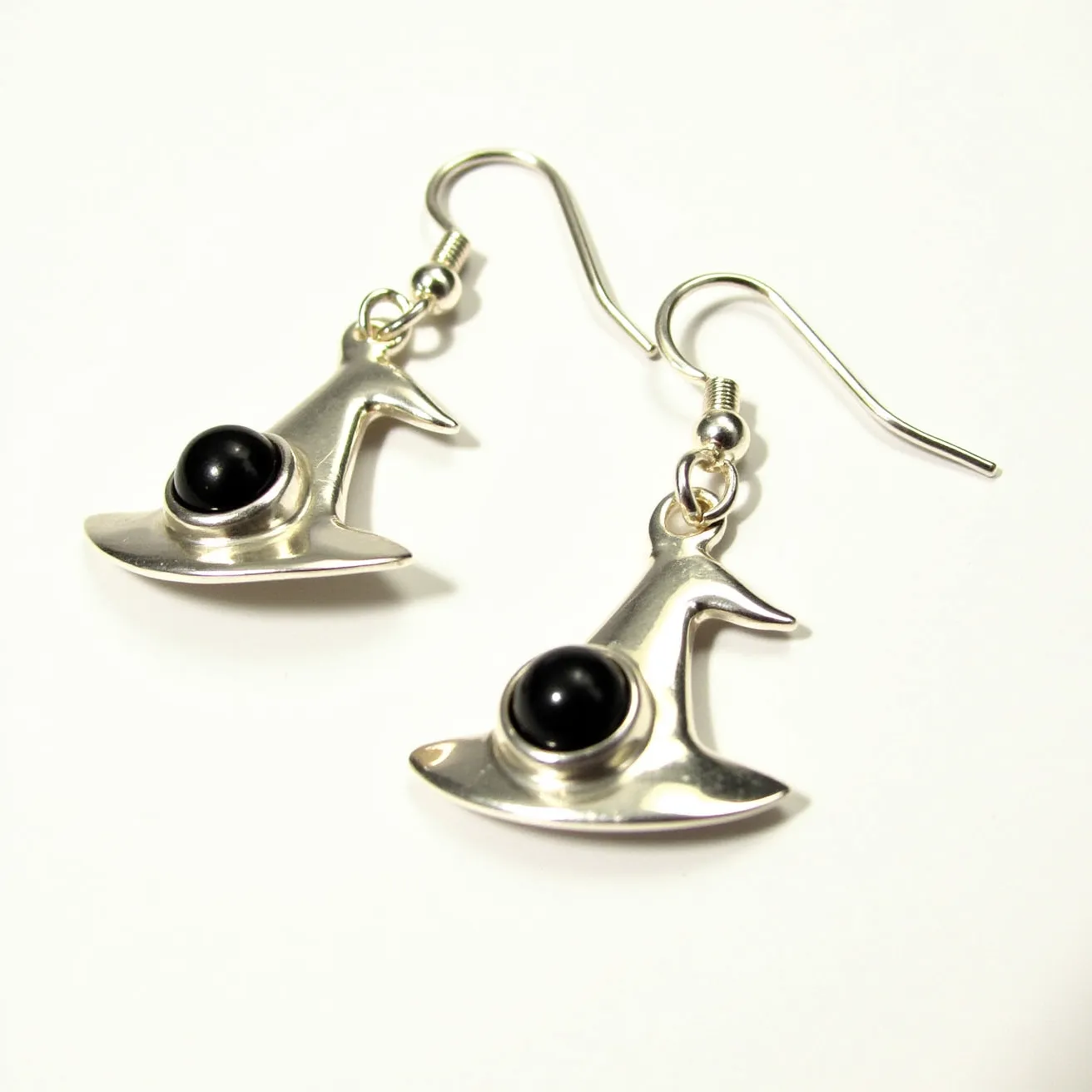 These Halloween earrings feature a charming witch hat design crafted from a shiny silver-colored metal. Each earring is adorned with a smooth, round black stone set prominently in the center of the hat, adding an eye-catching contrast. The stones are likely cabochon cut, smoothly polished without facets, enhancing their glossy appearance. The earrings are designed with a simple hook attachment, allowing for easy and secure wear. The overall aesthetic cleverly combines a festive theme with elegant elements, making them a delightful accessory for the Halloween season.