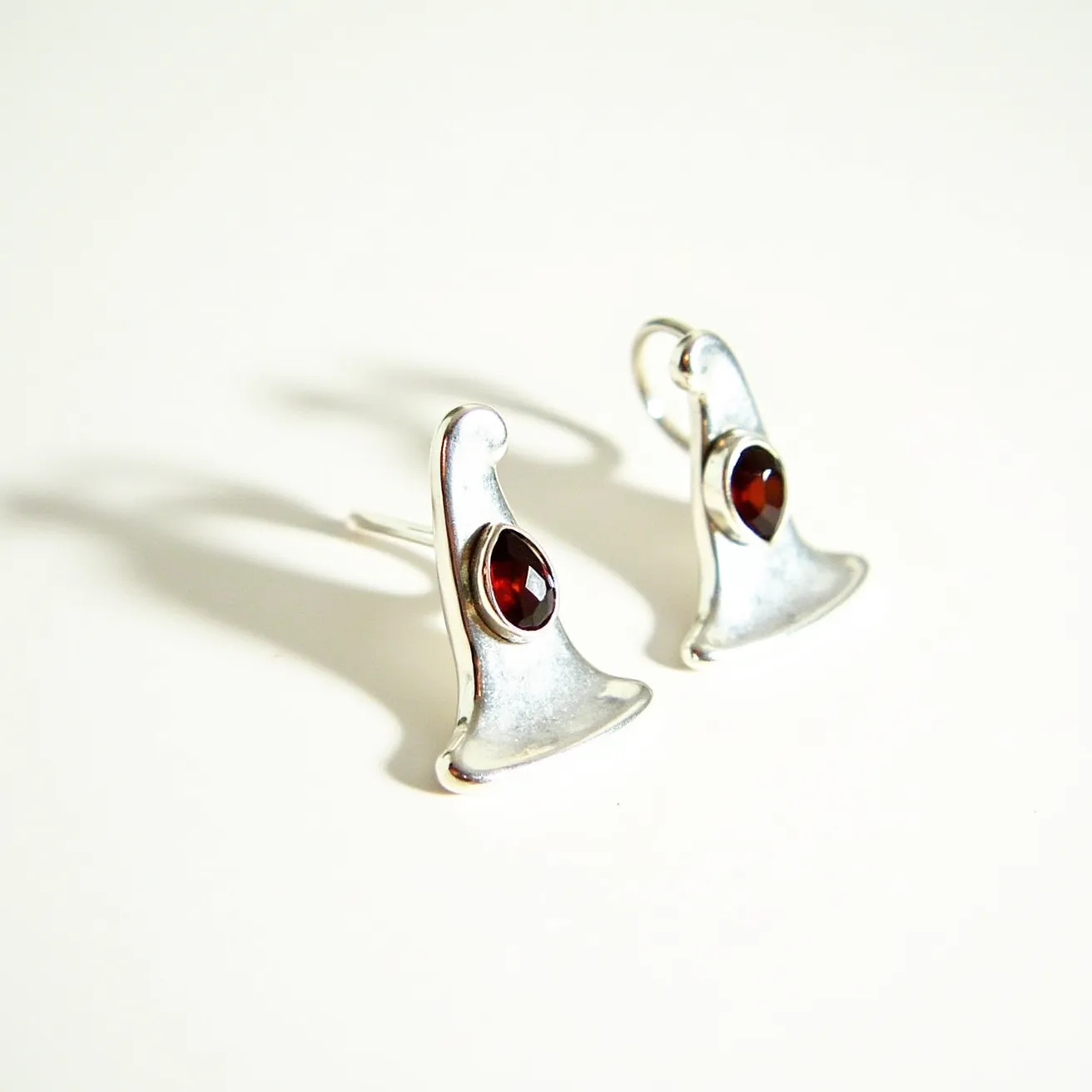 These Halloween earrings are crafted in the shape of whimsical witch hats, made from silver-toned metal. Each earring features a deep red, faceted gem, possibly garnet, with an oval cut, securely set in a bezel setting at the center of the hat design. The earrings are attached with traditional hooks, allowing them to dangle freely and capture attention with their playful yet elegant aesthetic.