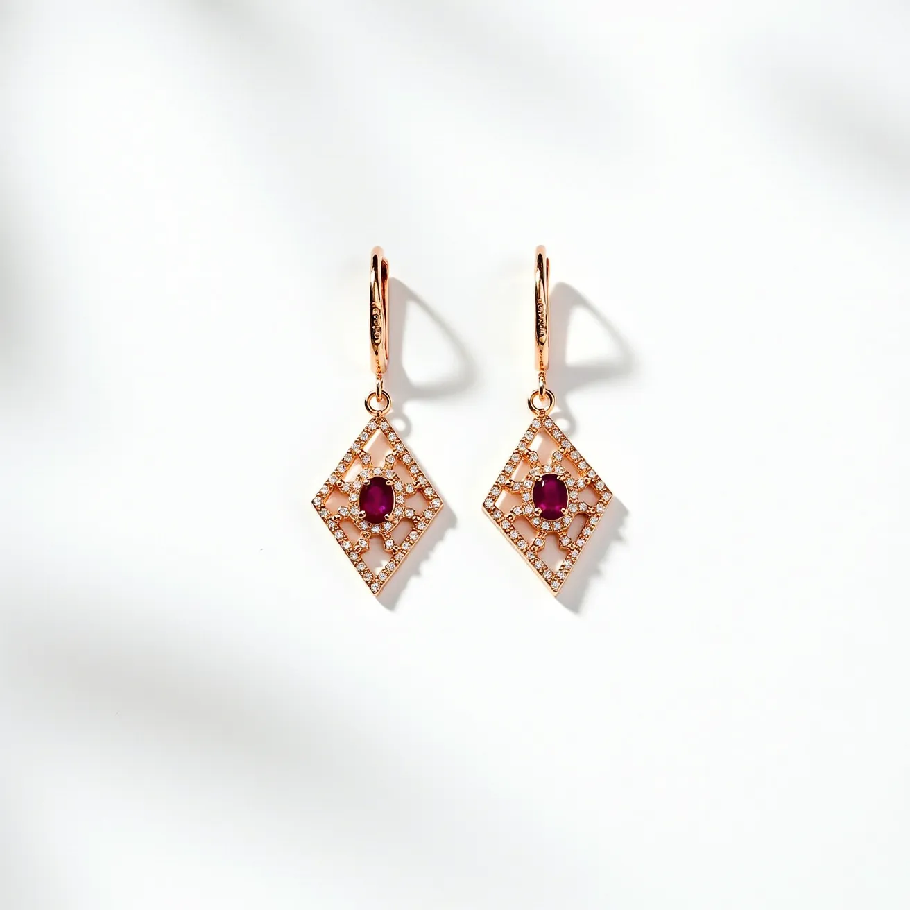 These hanging earrings feature a striking geometric diamond-shaped design crafted from what appears to be a rose gold metal. Each earring is adorned with a prominent oval-cut red gemstone, likely ruby, set prong-style at the center. The perimeter of the design and the intricate latticework are studded with numerous small, round, brilliant-cut clear stones, possibly diamonds, creating a sparkling contrast against the ruby. The earrings are suspended from a polished rose gold lever-back clasp, offering both security and elegance.