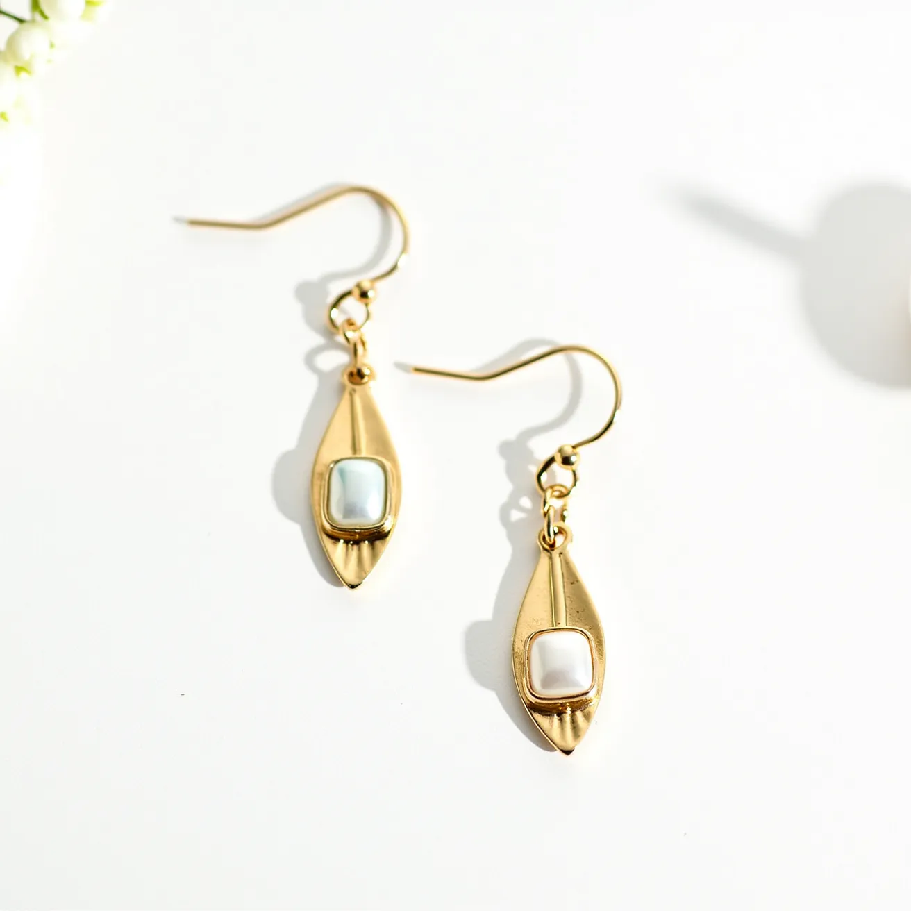 These hanging earrings feature a teardrop-shaped gold metal design with a modern and elegant charm. At the center of each earring is a square, bezel-set gemstone or pearl that appears to be a cabochon cut, providing a smooth, rounded surface with a lustrous finish. The stones are softly iridescent, adding a subtle touch of color and shine to the piece. The earrings are attached via a basic fishhook clasp, allowing them to dangle freely while being securely fastened to the ear. This combination of materials and the classic design create a timeless and versatile accessory suitable for various occasions.