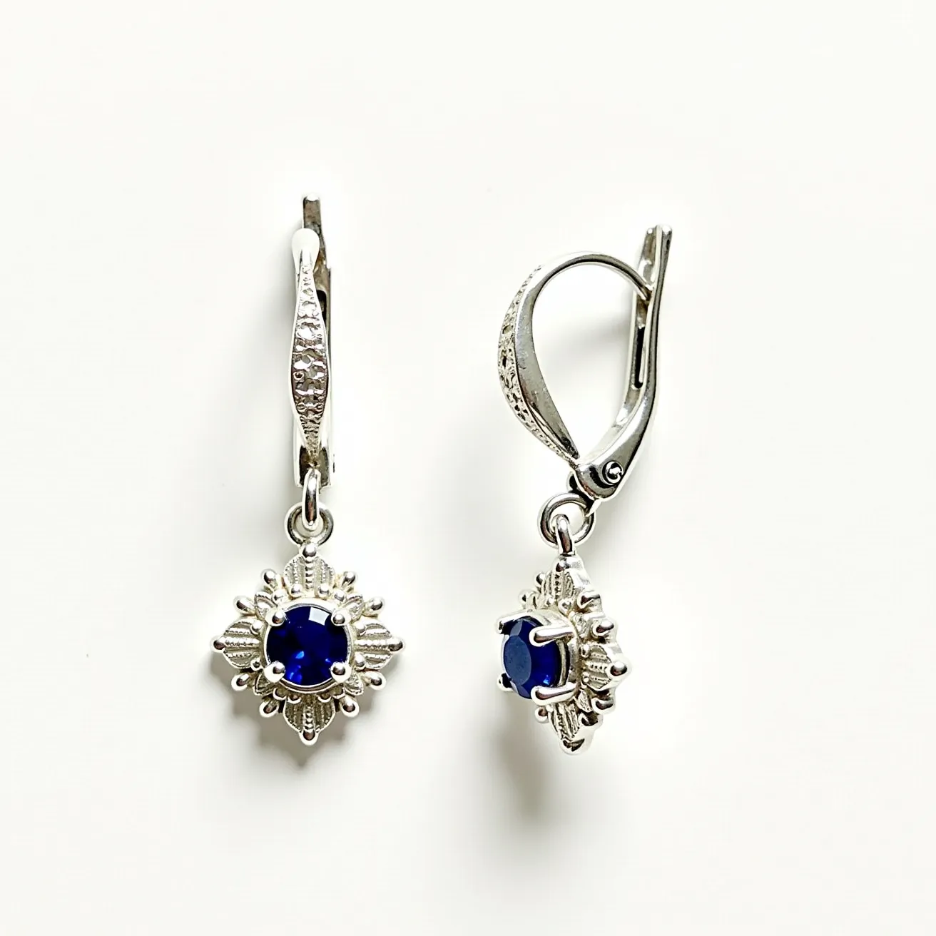 These pair of hanging earrings features a delicate design crafted from a silver-toned metal. At the center of each earring is a round, deep blue gemstone, possibly a sapphire, set in a prong setting that allows light to interact with the stone, enhancing its brilliance. Surrounding the central gem is an ornate, star-like embellishment that adds an intricate flourish to the overall design. The earrings are attached to the ear with lever-back clasps, providing secure and comfortable wear. The elegant combination of materials and the classic yet striking design make these earrings a sophisticated accessory.