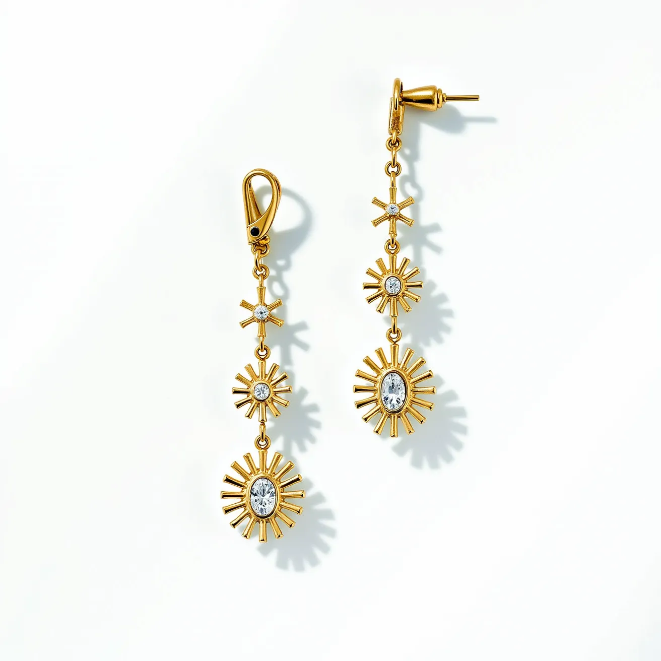 These hanging earrings feature a striking design crafted from a gold-toned metal, accentuating the elegant and contemporary aesthetic. Each earring consists of a series of three starburst motifs descending vertically, with the central and largest bursts prominently showcasing a clear, faceted oval gemstone set securely in a bezel setting. The smaller bursts are adorned with round, clear gemstones, providing a coherent and sparkling look. The earrings feature two types of attachments: a fishhook clasp on one and a post with a butterfly backing on the other, both ensuring secure and comfortable wear. The combination of materials and detailed craftsmanship highlights the intricate and radiant appearance of these earrings.