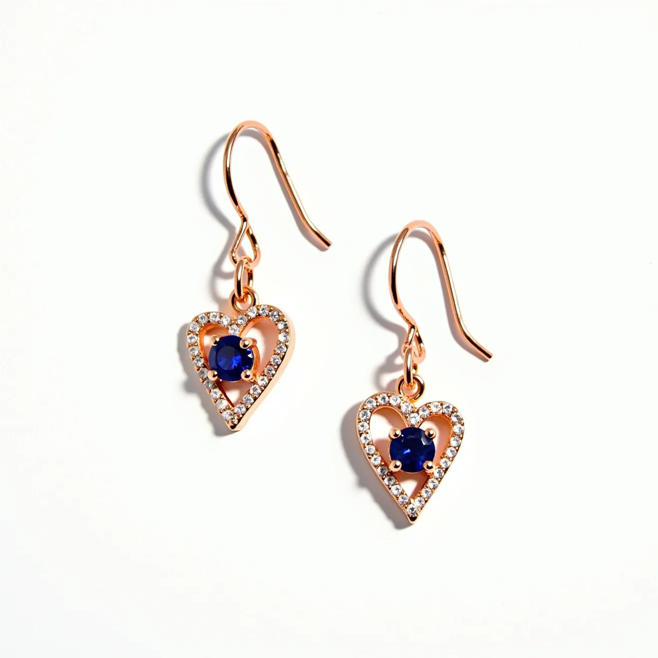 These hanging earrings feature a delicate and elegant design with a heart-shaped motif crafted from rose gold metal. Each earring is adorned with a central sapphire-like gem, round in shape and held in a secure four-prong setting. Surrounding the central stone, there is a halo of small, clear cubic zirconia or diamond-like stones set closely together, adding a shimmering border to the design. The earrings are suspended by a classic shepherd hook clasp, allowing for easy wear and a gentle sway with movement.