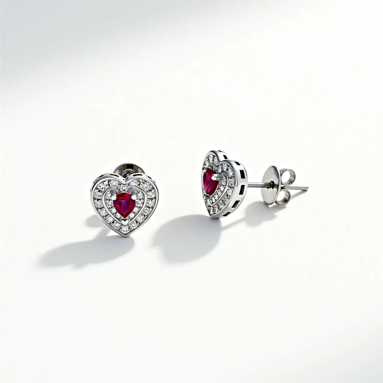 These heart diamond earrings feature an intricate design that combines white and red gemstones. The central focus is a heart-shaped red gemstone, possibly a ruby, set prominently at the center of each earring. Surrounding the red gemstone is a halo of smaller round white diamonds that enhance the brilliance and elegance of the design. The setting is likely made of a silvery metal, possibly white gold or platinum, which adds to the luxurious appearance. The earrings are secured with a classic post and butterfly back clasp, ensuring a secure fit when worn.