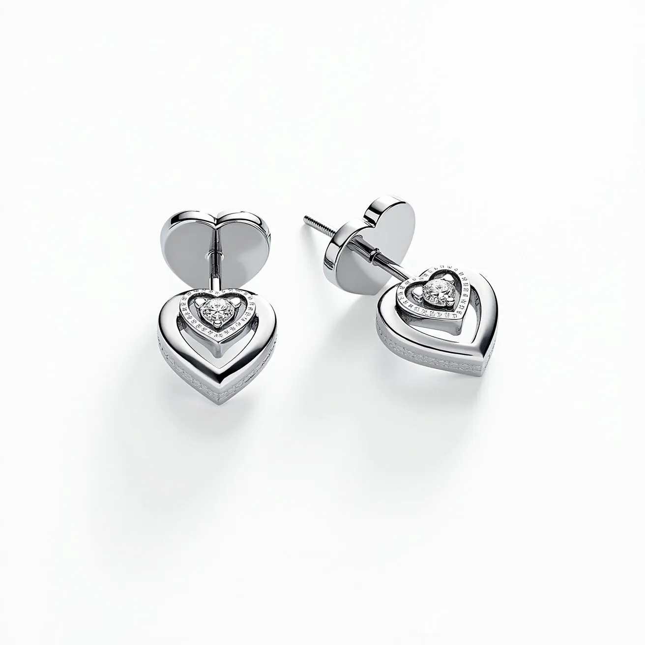 These heart diamond earrings feature a central heart-shaped diamond in each piece, intricately set within a stylish heart-shaped mount. The diamonds are bezel-set, emphasizing their brilliance and sparkle. The mount is likely crafted from a polished precious metal, such as white gold or platinum, which complements the diamonds' clarity and enhances their reflective quality. Surrounding the central diamond is a fine line of accent stones that add to the earrings' elegance. The earrings utilize a screw-back attachment, ensuring a secure and comfortable fit for the wearer.