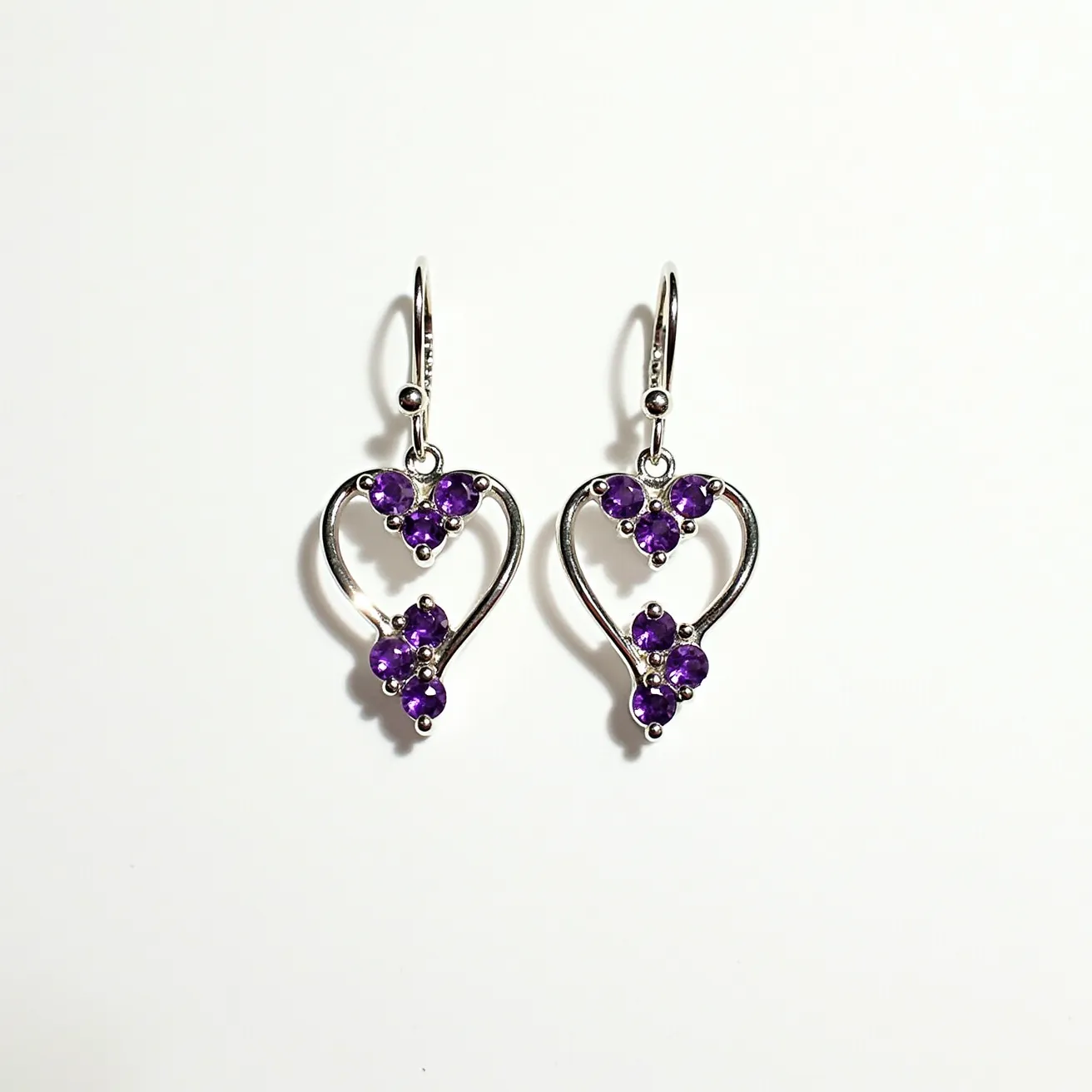 These heart earrings feature an elegant design crafted from a metallic material, possibly silver, forming an open-heart shape. The earrings are adorned with purple gemstones, likely amethysts, set in a prong setting. Each earring contains multiple round-cut stones artistically arranged to complement the heart outline. The earrings are equipped with a fishhook attachment, providing a secure yet easy-to-use clasp system. The combination of the metallic sheen and vibrant gemstones creates a striking and sophisticated appearance.