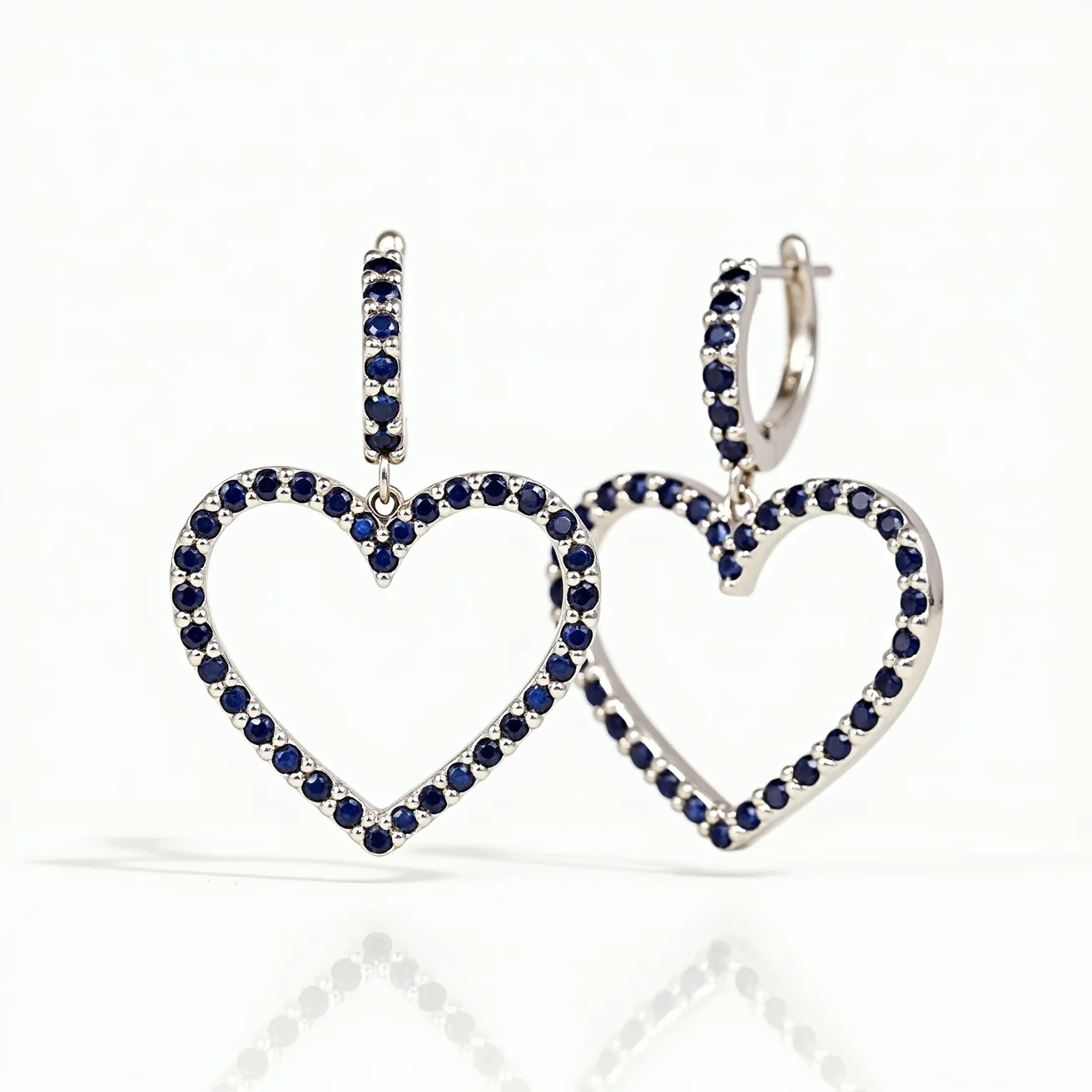 These heart earrings feature a delicate design crafted from a metallic, likely silver or white gold, framework. Each heart earring is adorned with a series of small, round dark blue stones, which are presumably sapphires. The stones are uniformly set in a prong setting, allowing them to be securely attached while maximizing their visibility and sparkle. The earrings connect through a small hoop, which also features the same blue stones, and likely employs a latch-back clasp for securing them comfortably on the ear. This design combines elegance and sophistication, highlighted by the vibrant color of the gemstones against the cool-toned metal.