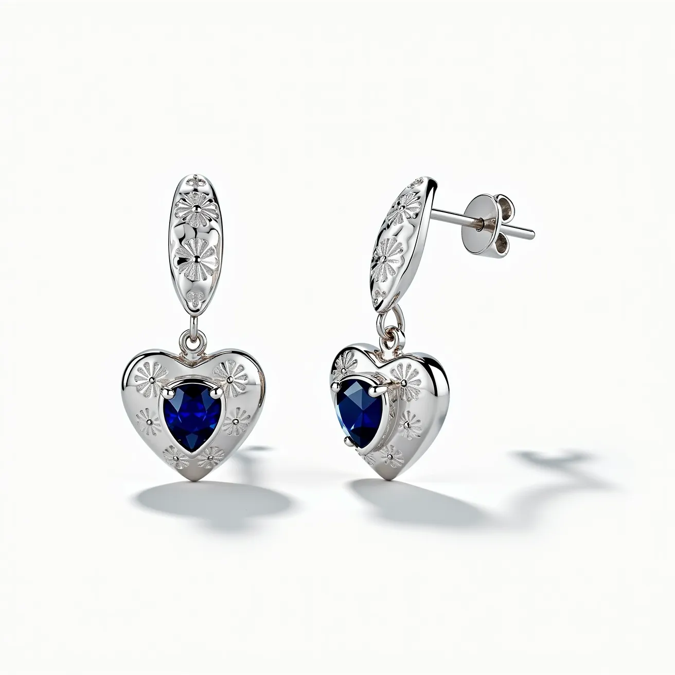 These heart earrings feature a striking design with a deep blue, heart-shaped gemstone, possibly a sapphire, set prominently in each earring. The stones are faceted to enhance their sparkle and are secured in a bezel setting. The earrings are crafted from a metal that appears to be silver, with intricate floral engravings decorating the surface, adding elegance and texture. The attachment consists of a stud and post securing system, ensuring they sit comfortably and securely when worn. The overall aesthetic is both classic and sophisticated, blending the romantic heart motif with timeless materials and design elements.